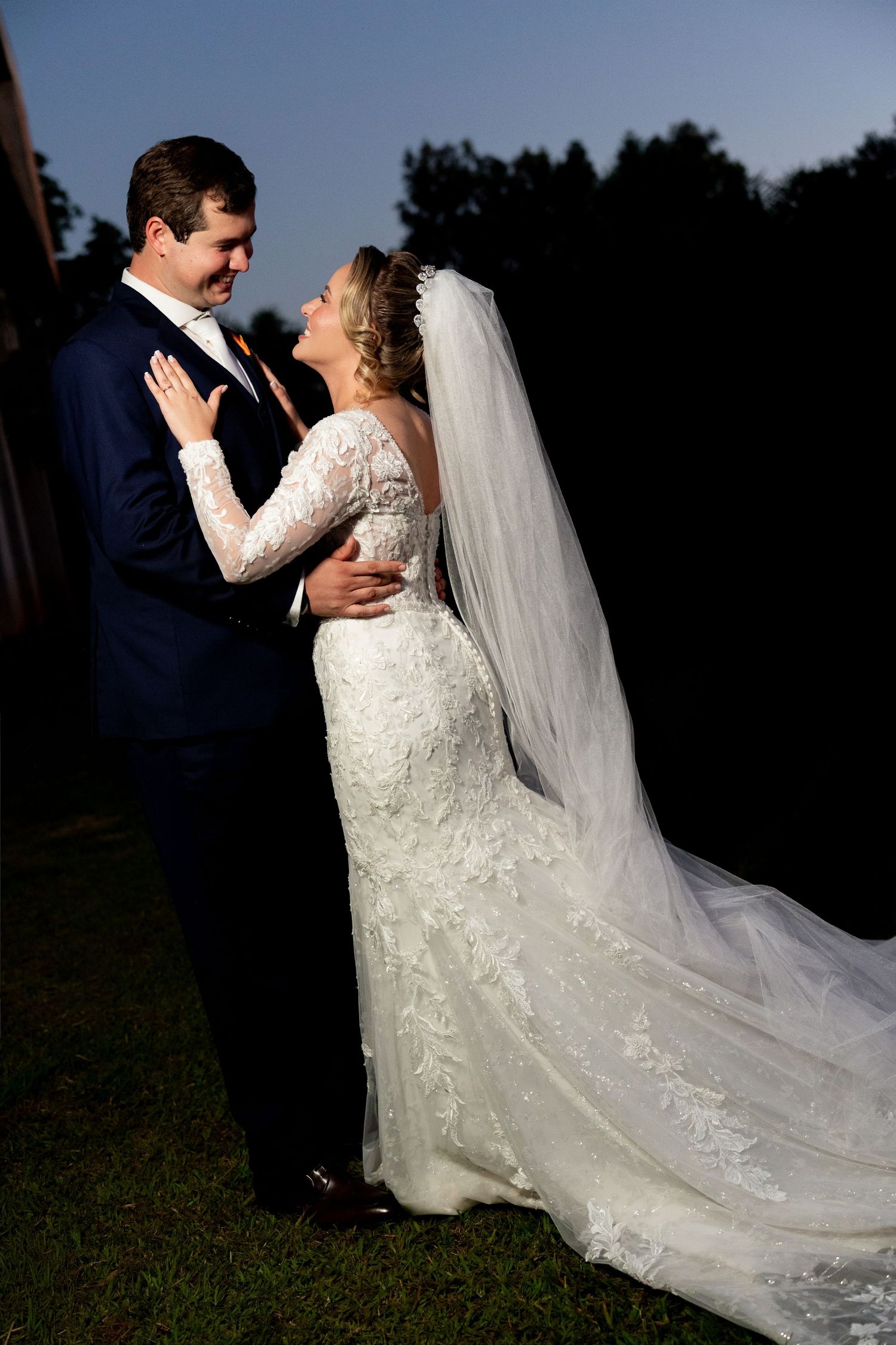 Bride in lace gown and veil gazes at groom in navy suit during romantic outdoor wedding portrait at sunset.