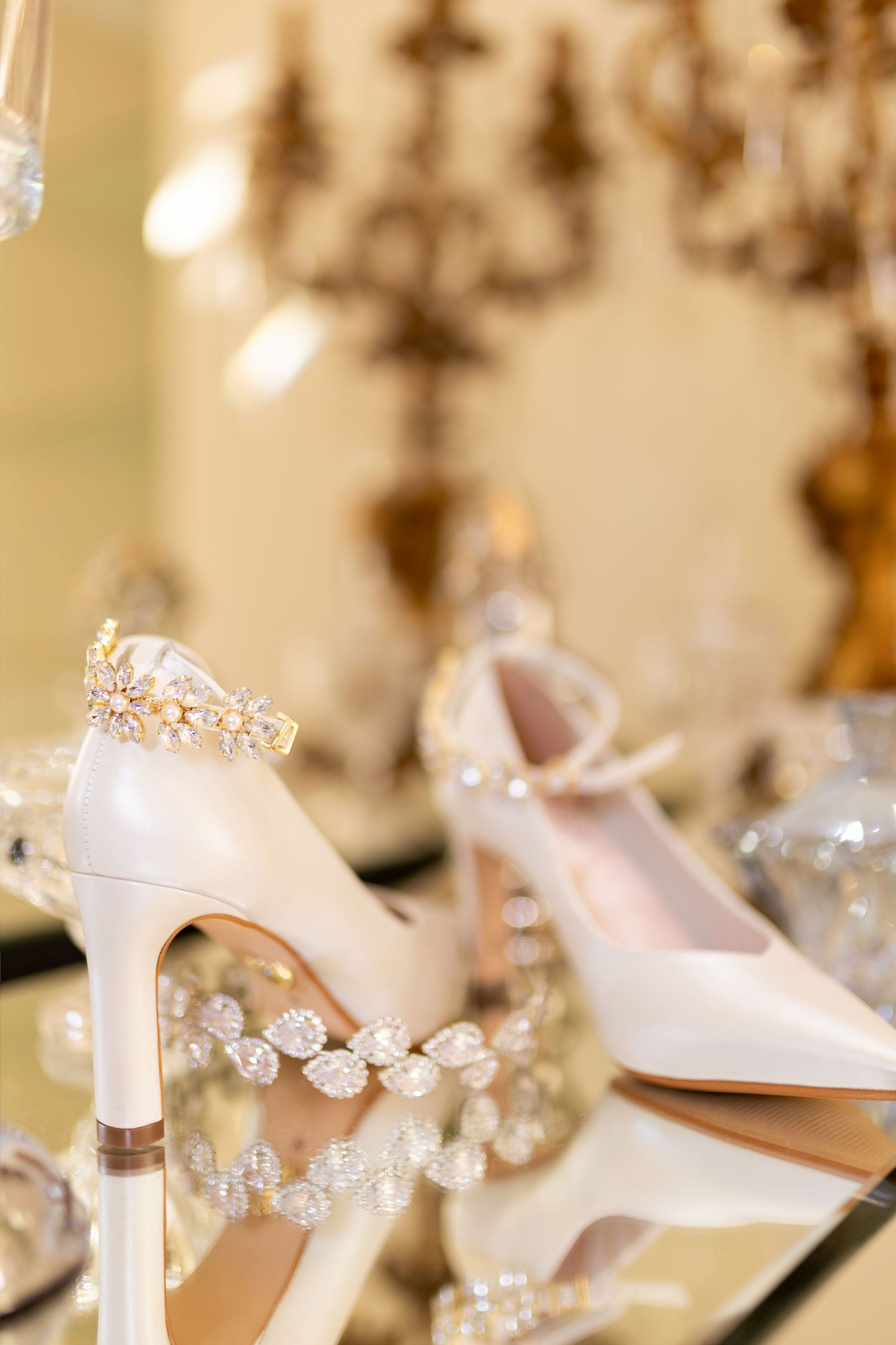 Elegant white bridal heels with crystal embellishments displayed on a reflective surface with candelabra in background.