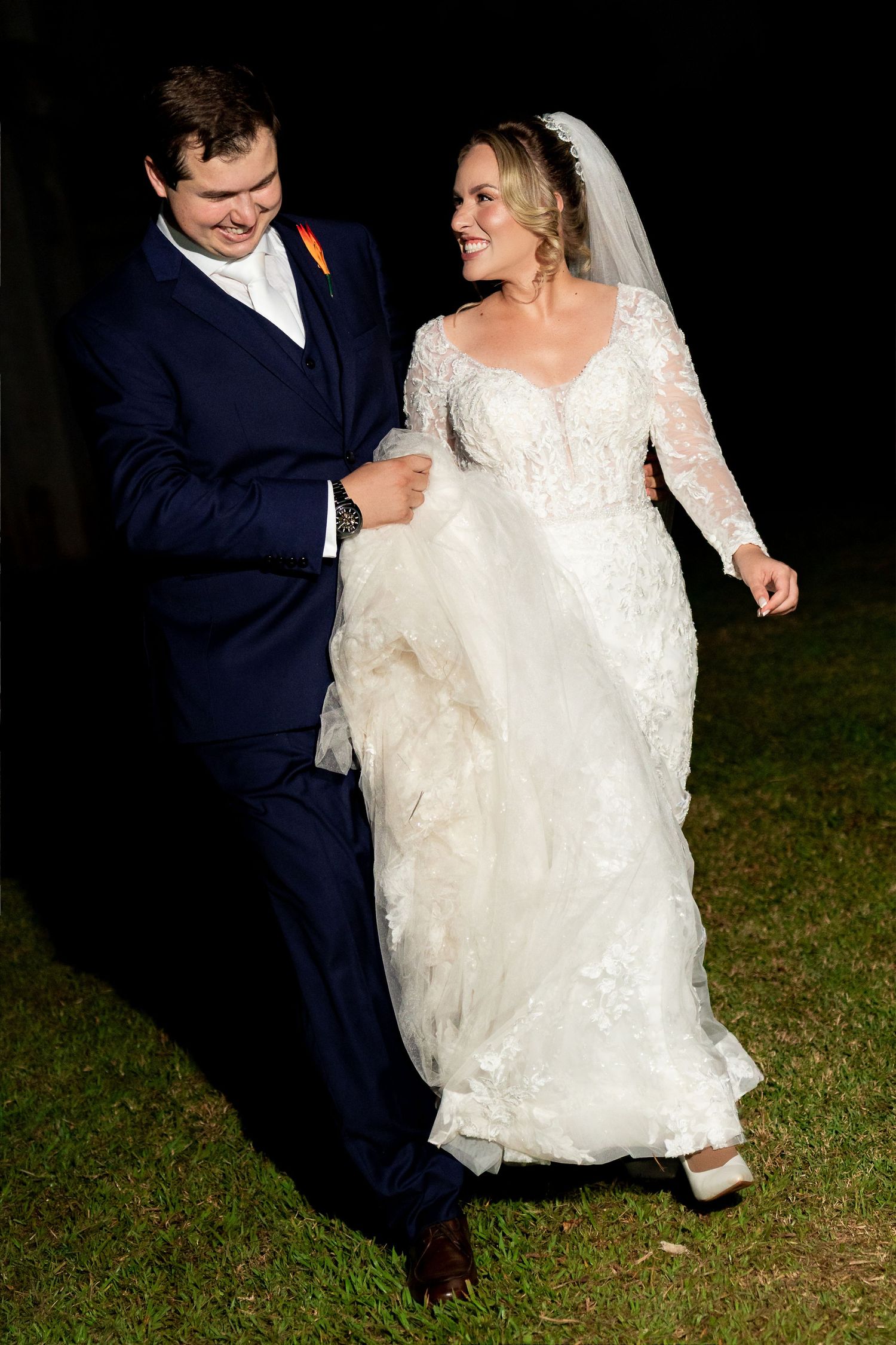 Bride in lace gown and groom in navy suit walk together smiling at night during outdoor wedding celebration.