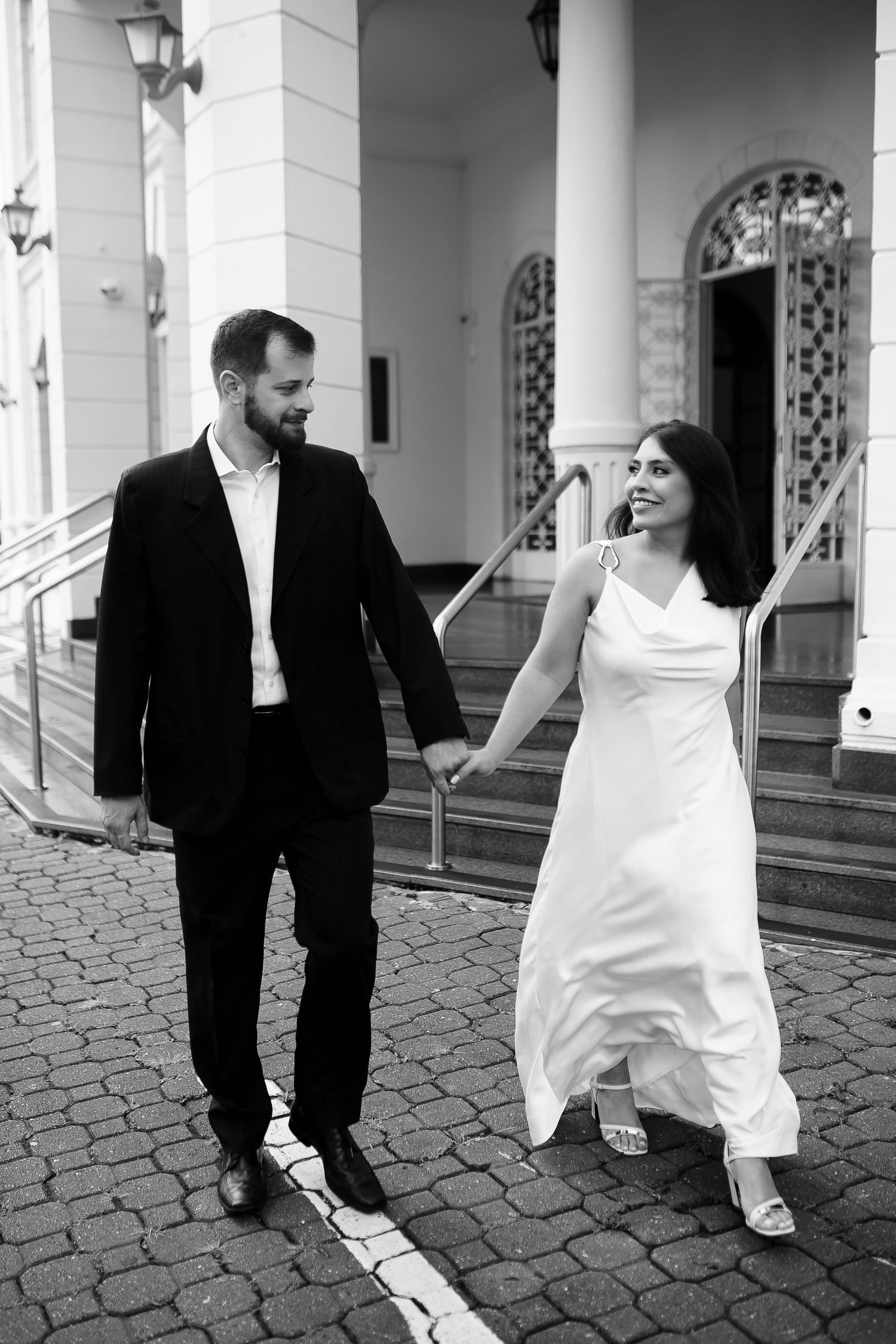 Black and white photo of a smiling bride in white gown and groom in suit holding hands walking outside a building.
