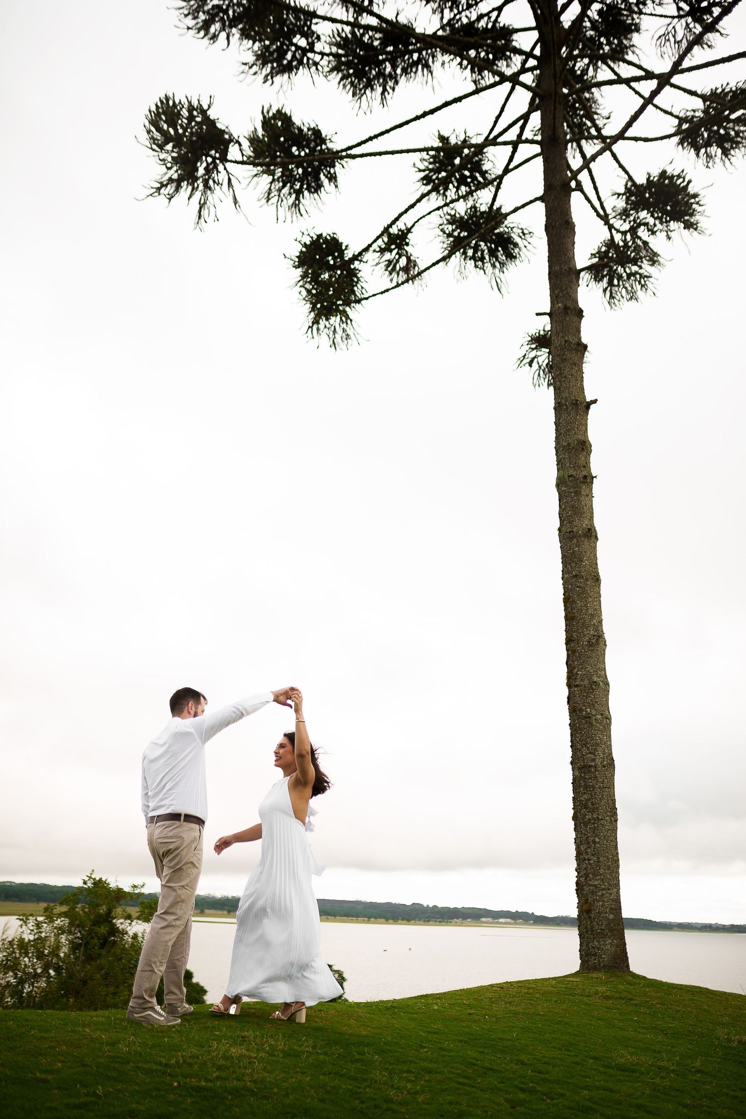 Couple dancing outdoors near a tall tree by a lake, bride in white dress being twirled by groom in light attire.