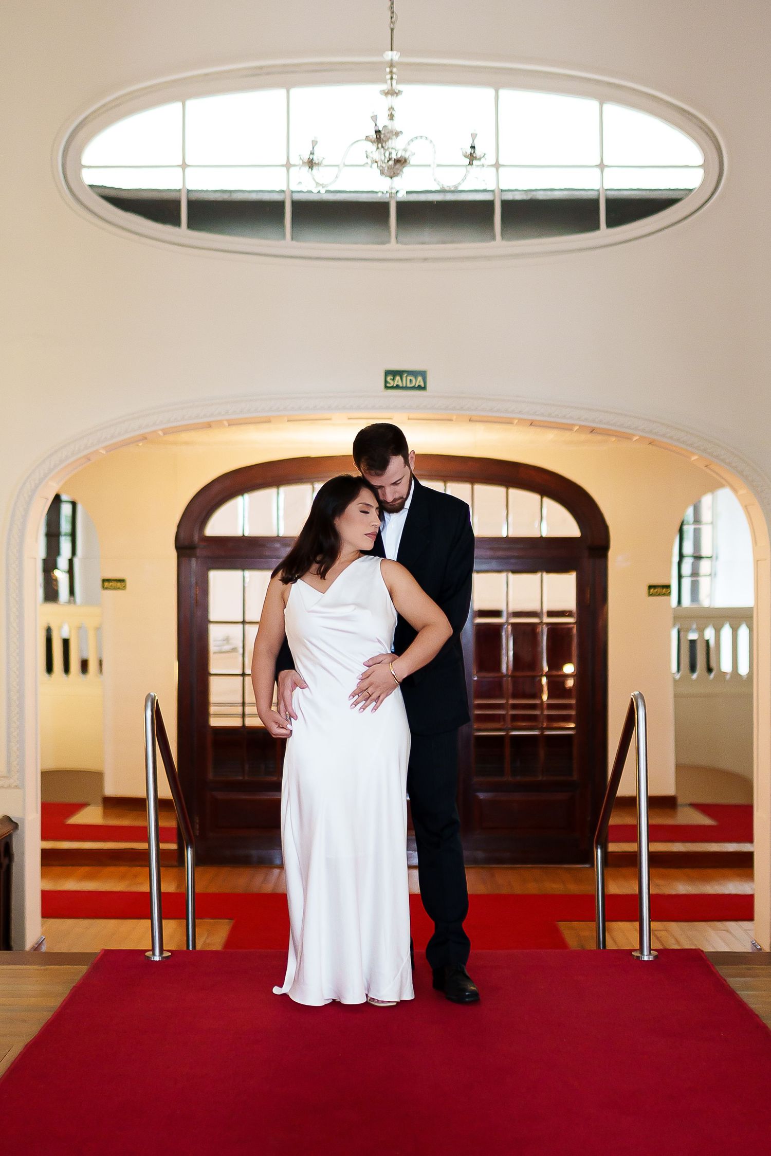 Elegant bride and groom pose on red carpet steps in a grand hall with arched wooden doors.