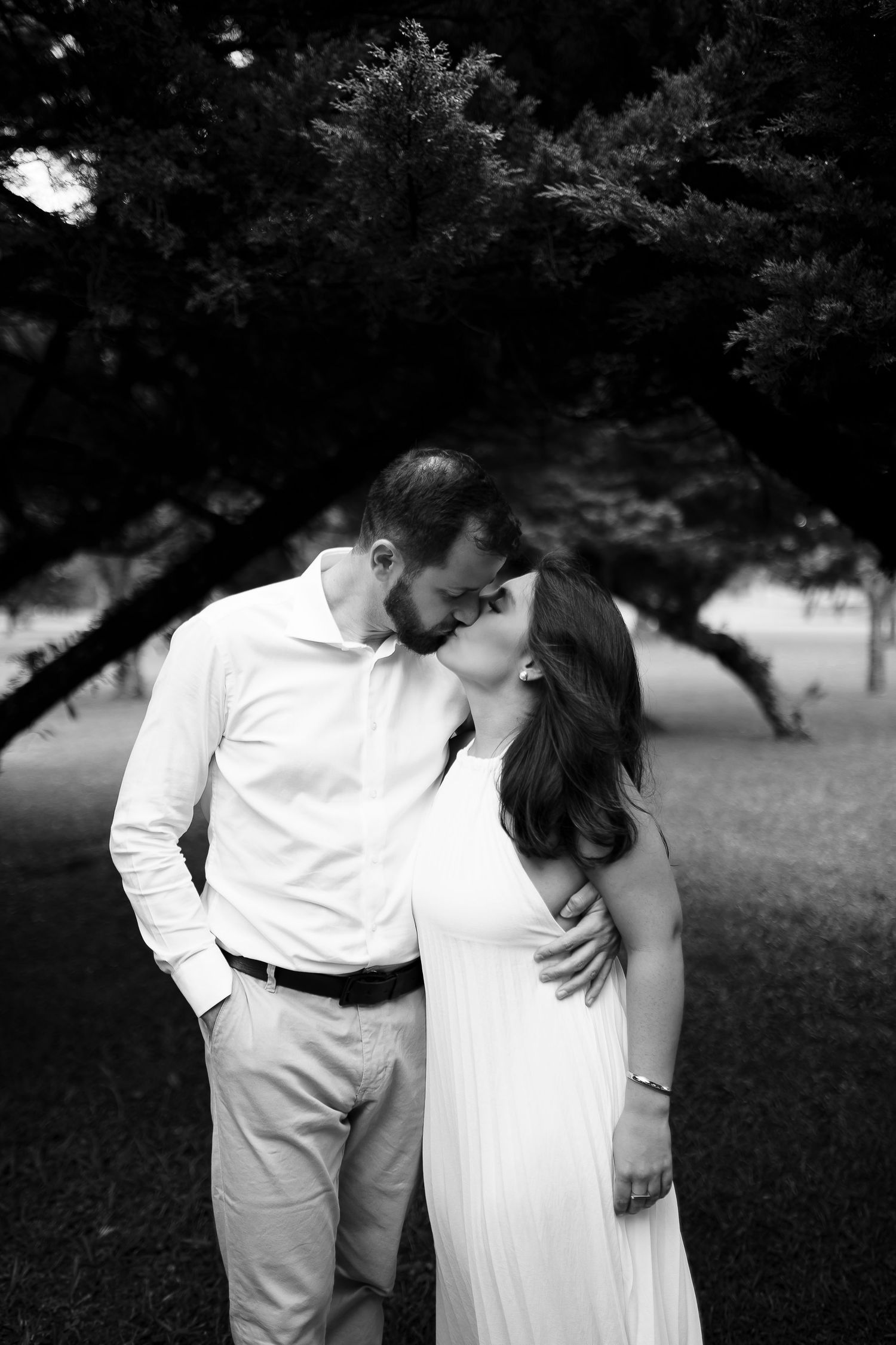 Black and white photo of a couple kissing outdoors under a large tree, woman in white dress, man in white shirt.