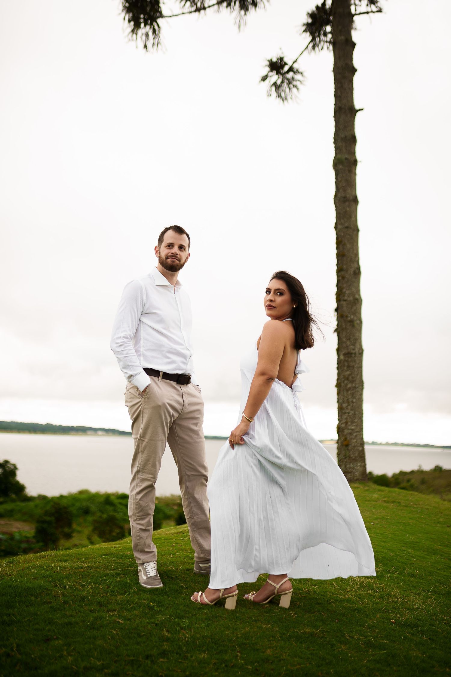 Couple in white outfits standing on green lawn near tall tree with water view in background.