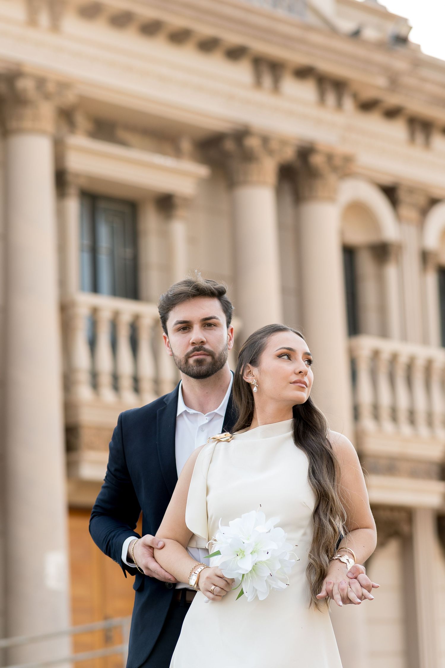 Elegant bride in white dress holding bouquet with groom in navy suit posing before a classical building.