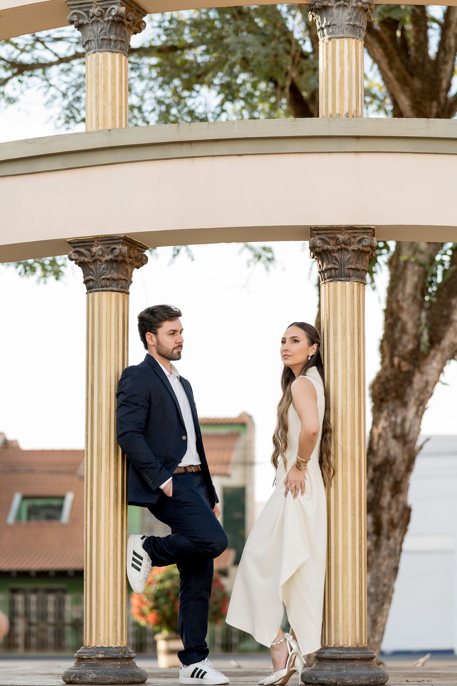 Couple posing under elegant gold columns outdoors, man in navy suit and woman in white gown with long flowing hair.