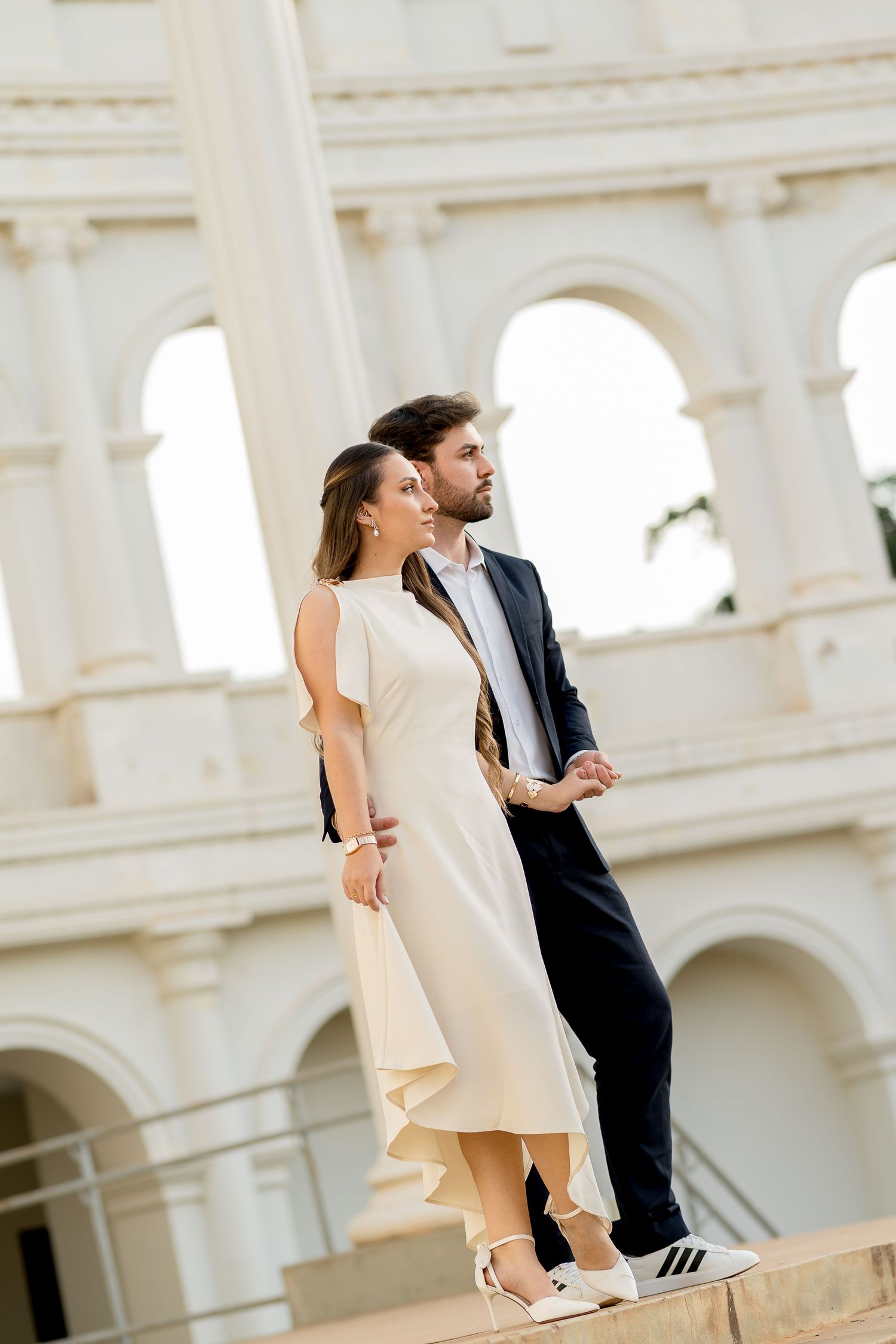 Elegant couple in formal attire posing in front of a grand white arched building, woman in cream dress, man in navy suit.