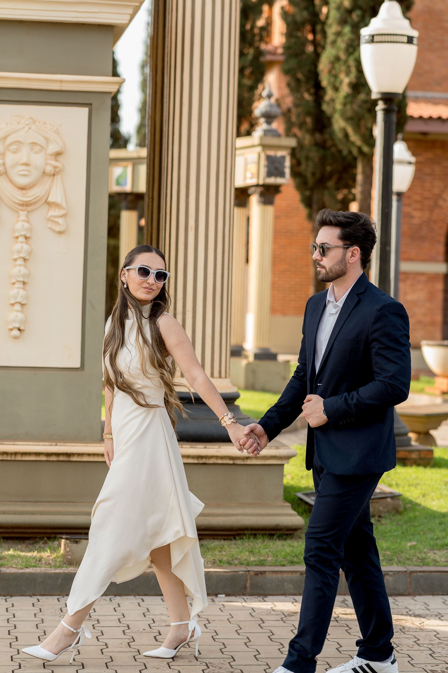 Elegant couple holding hands walking outdoors, woman in white dress and sunglasses, man in dark suit.