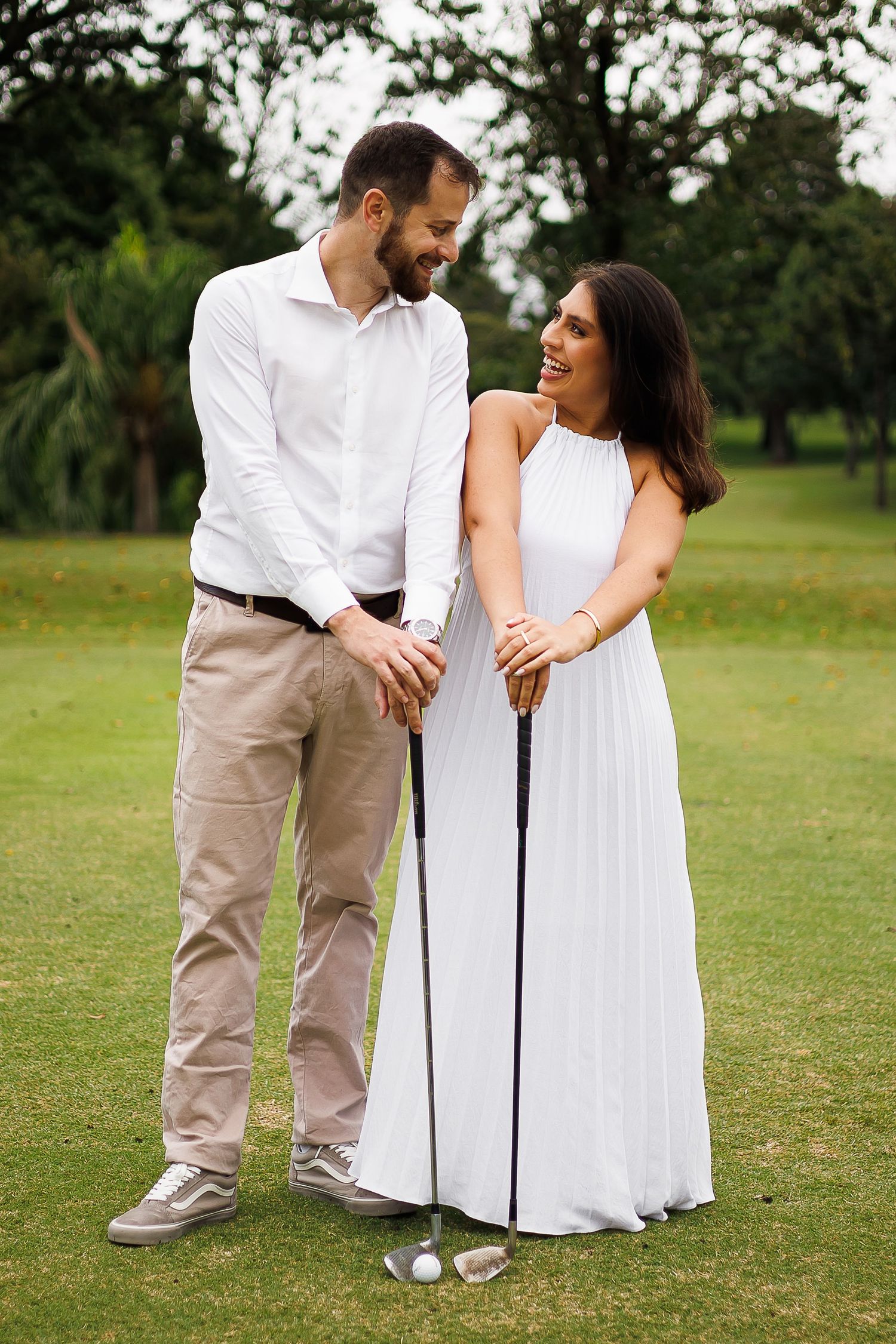 Couple smiling together on a golf course, woman in white dress holding a golf club on a green fairway.
