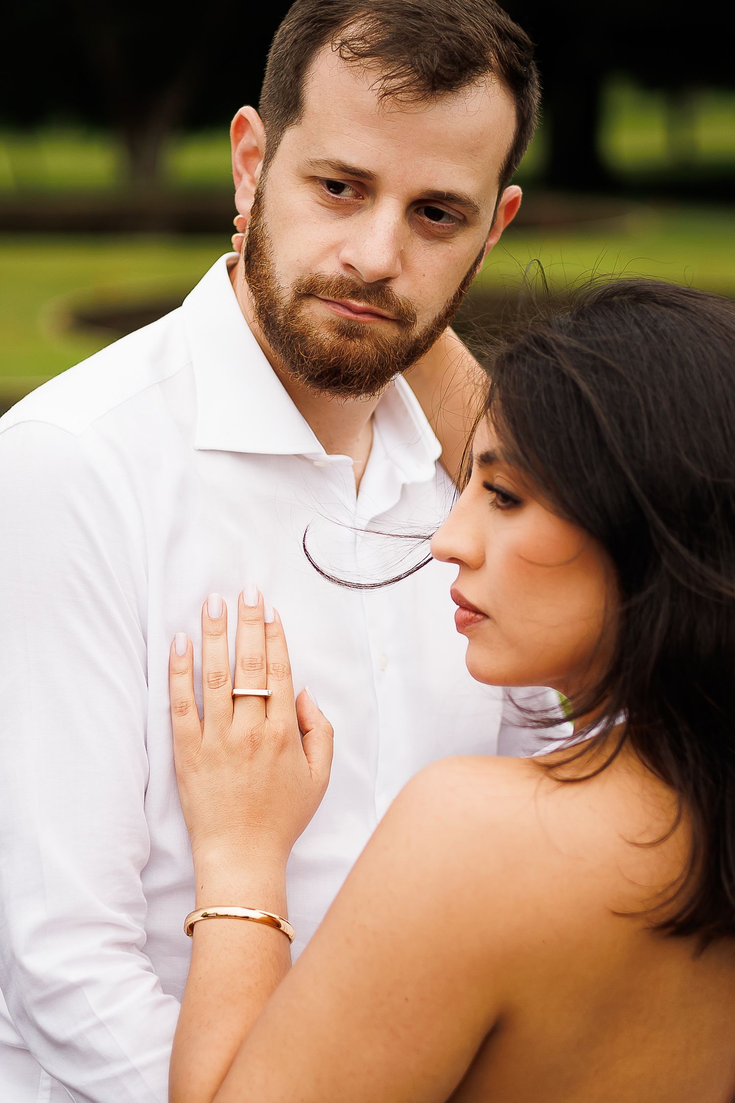Romantic couple embracing outdoors, woman with wedding ring resting hand on man's chest in a garden setting.