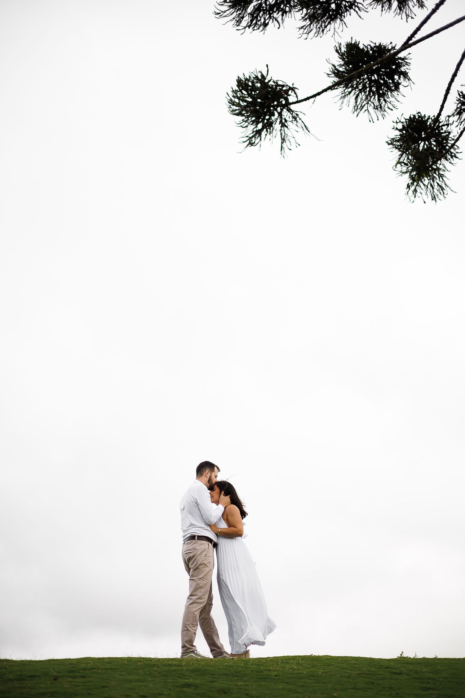 Couple embracing on green hill in white wedding attire under pine tree branches against overcast sky.