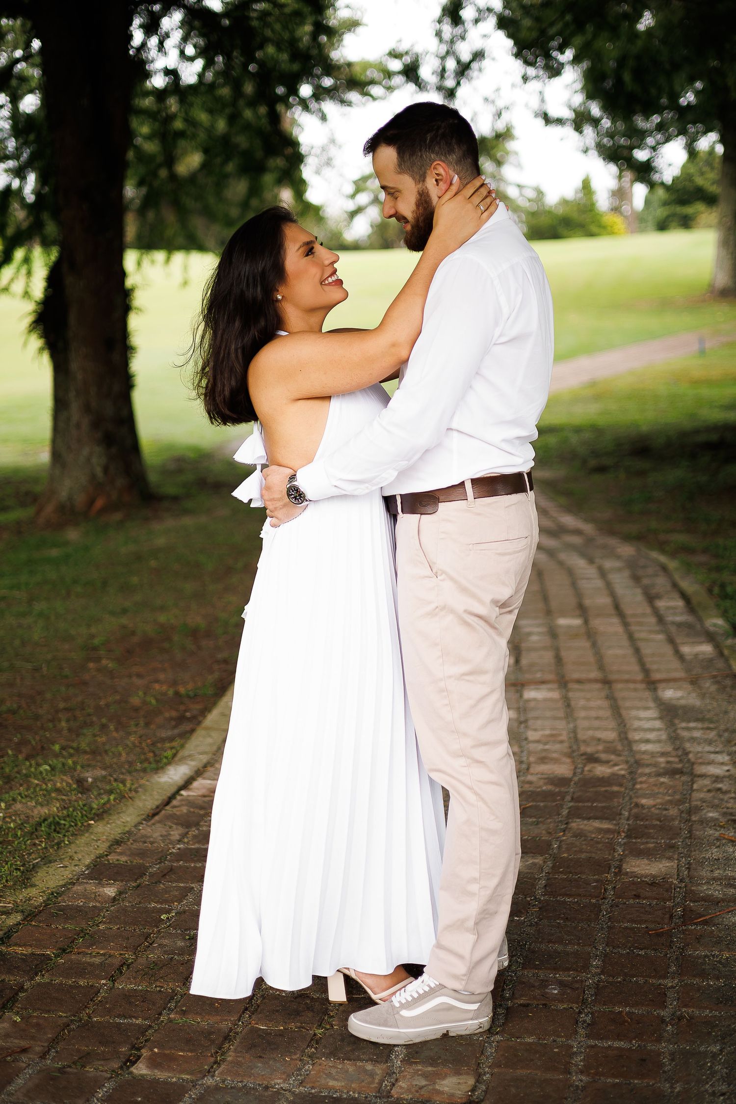A couple in white and beige embraces on a brick path in a lush green park, sharing a romantic moment.