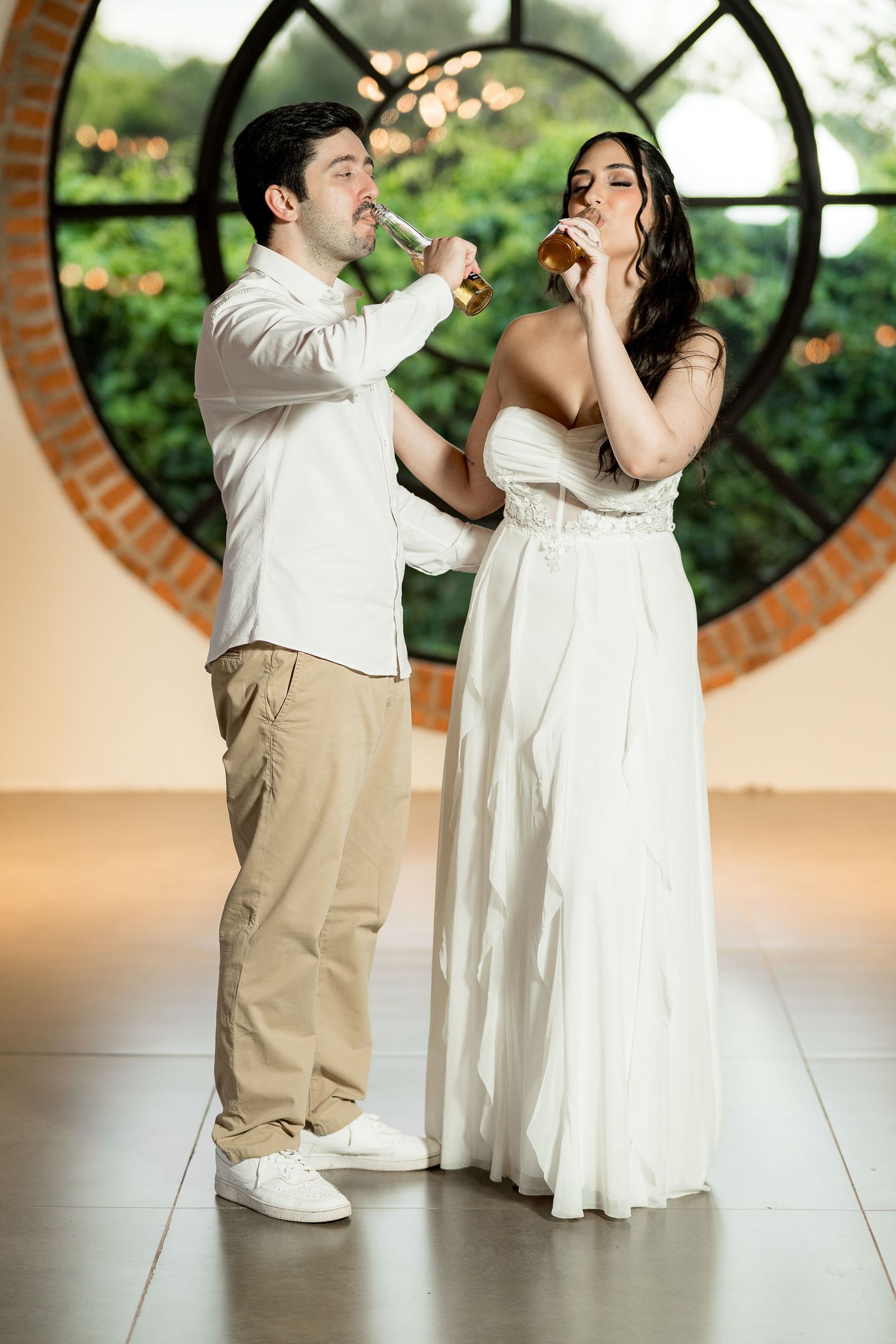 Bride and groom drinking beer bottles together in front of a round window at their wedding reception.