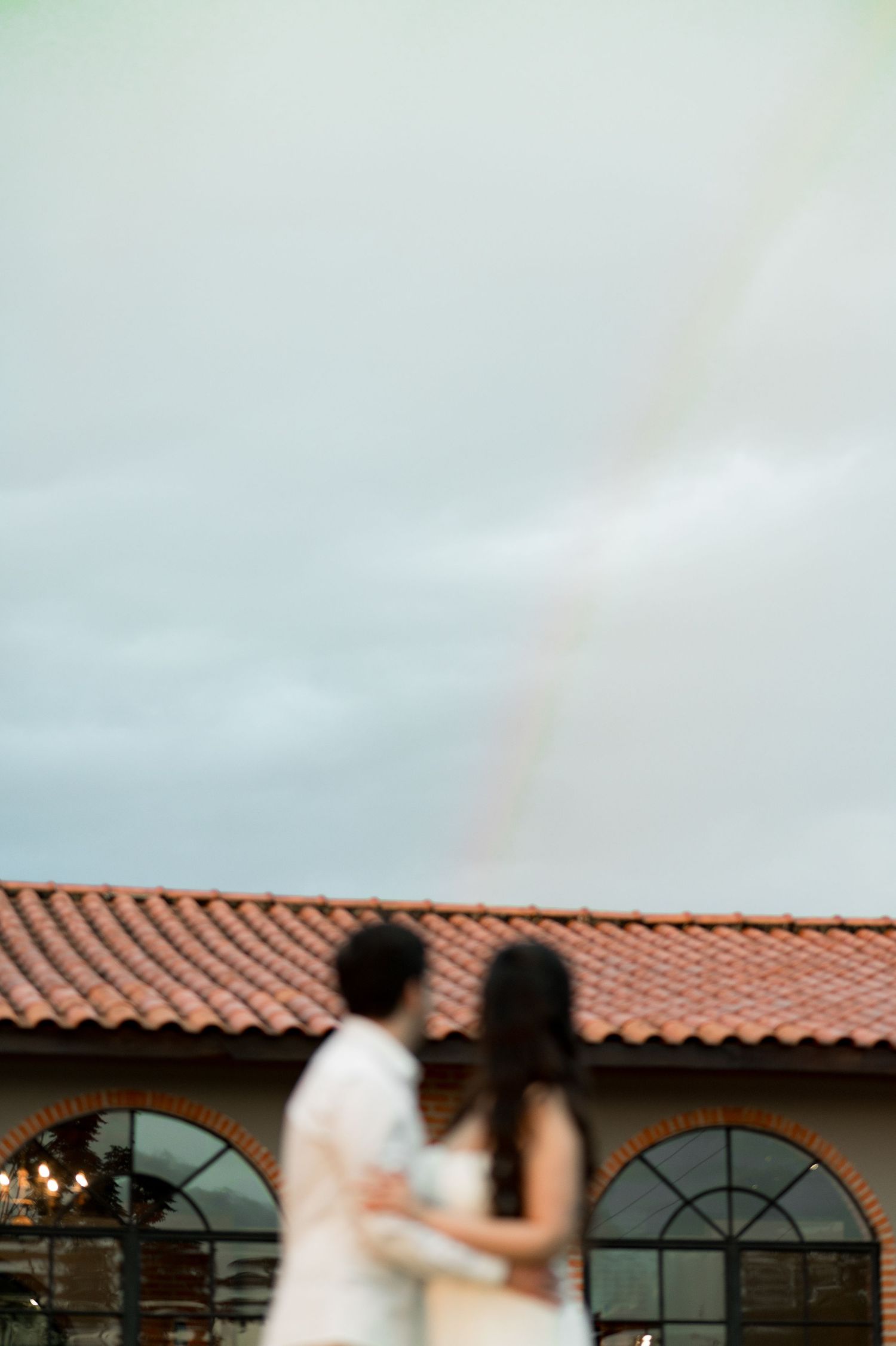Blurred couple embracing outdoors with a rainbow over a terracotta roof building in the background.