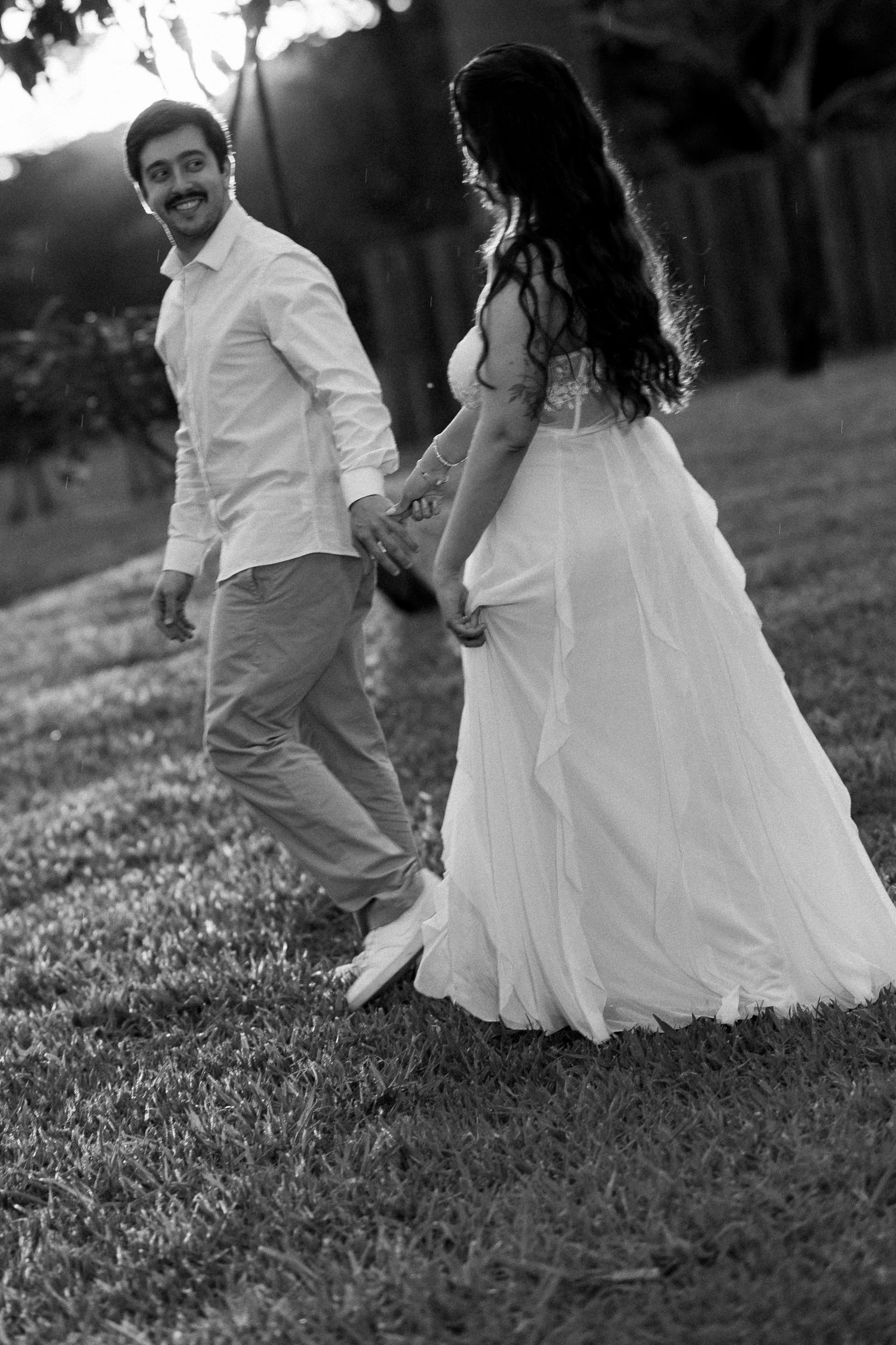 Black and white photo of a smiling groom leading his bride by the hand across a grassy lawn at sunset.