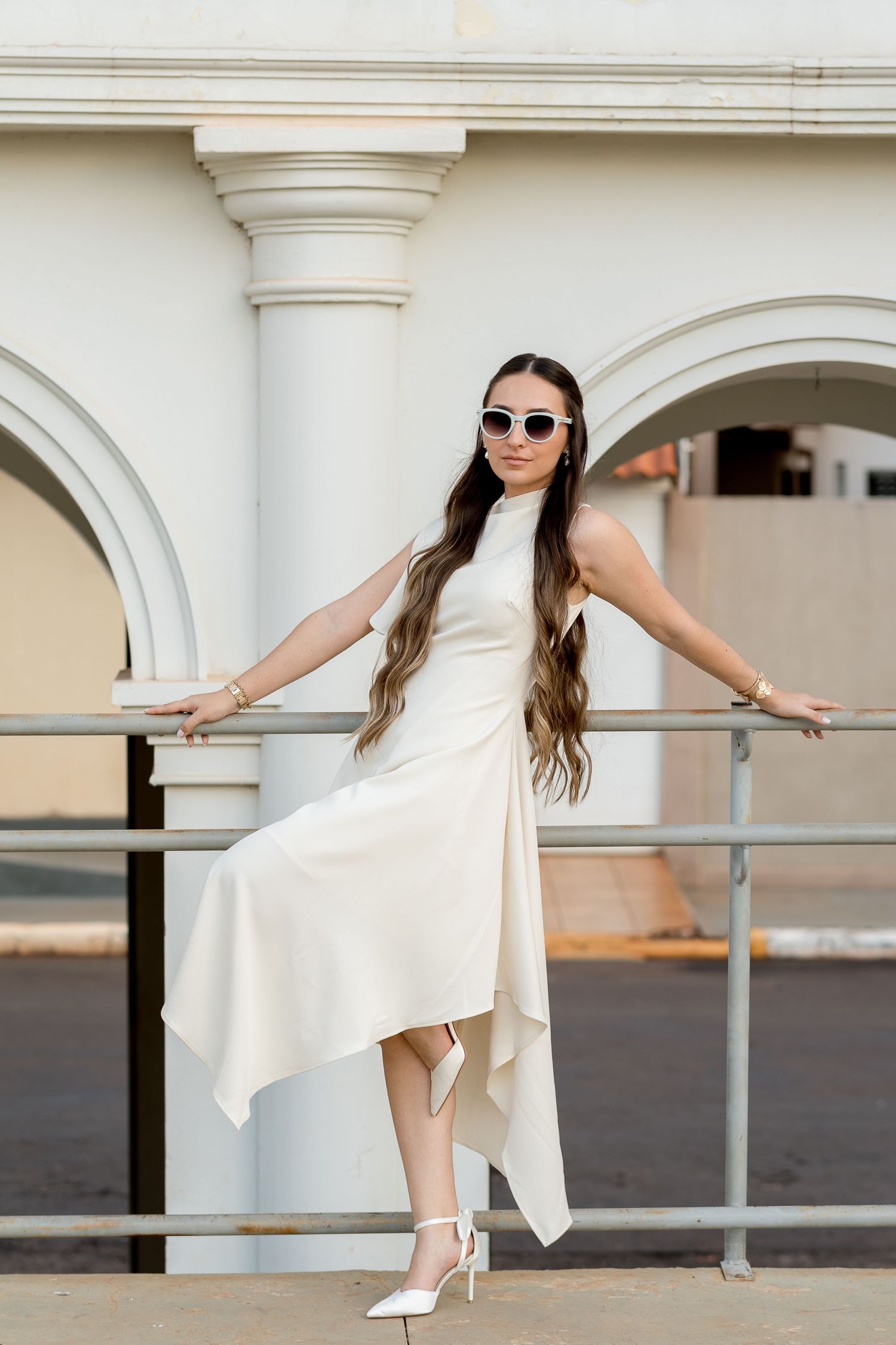 Elegant woman in white asymmetric dress and sunglasses posing by metal railing near white arched columns.