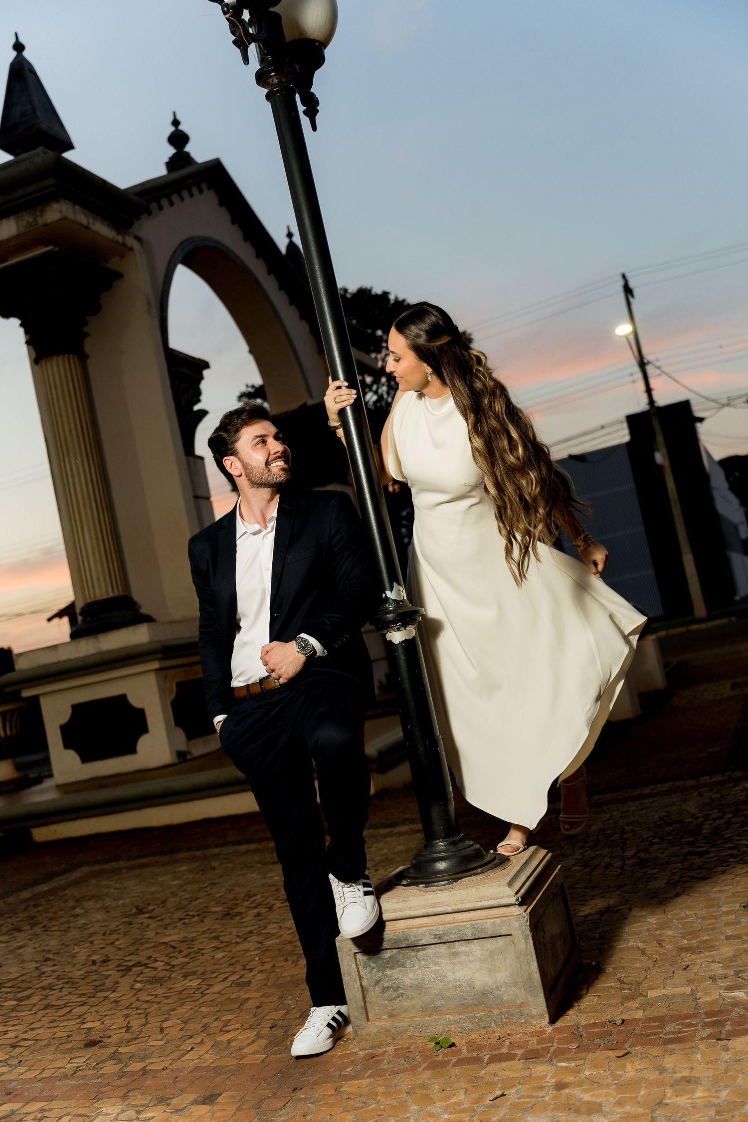 Couple poses at sunset near ornate lamppost, woman in white dress standing, man in suit sitting on cobblestone plaza.