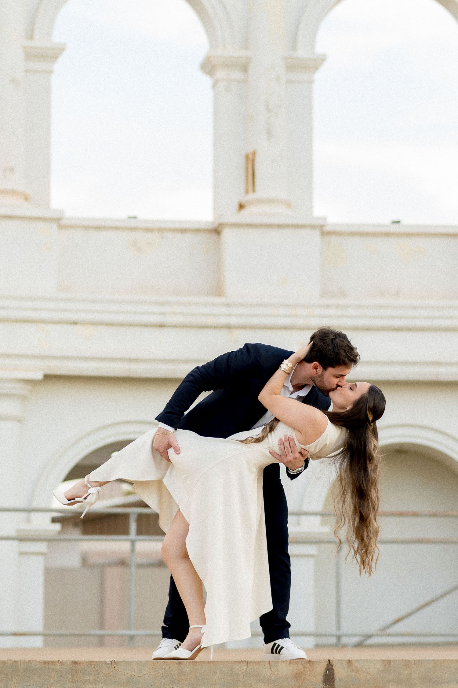 A groom dips his bride for a romantic kiss in front of elegant white arched columns.