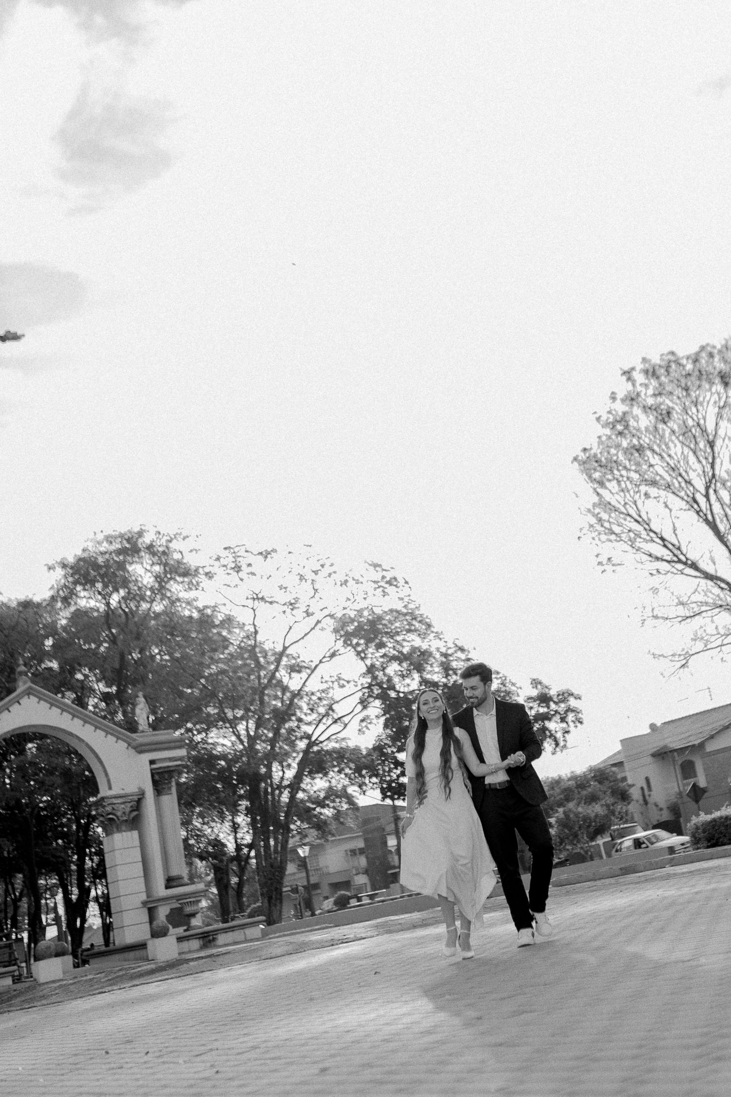 Black and white photo of a couple in wedding attire walking together on a paved path with trees and an arch in background.