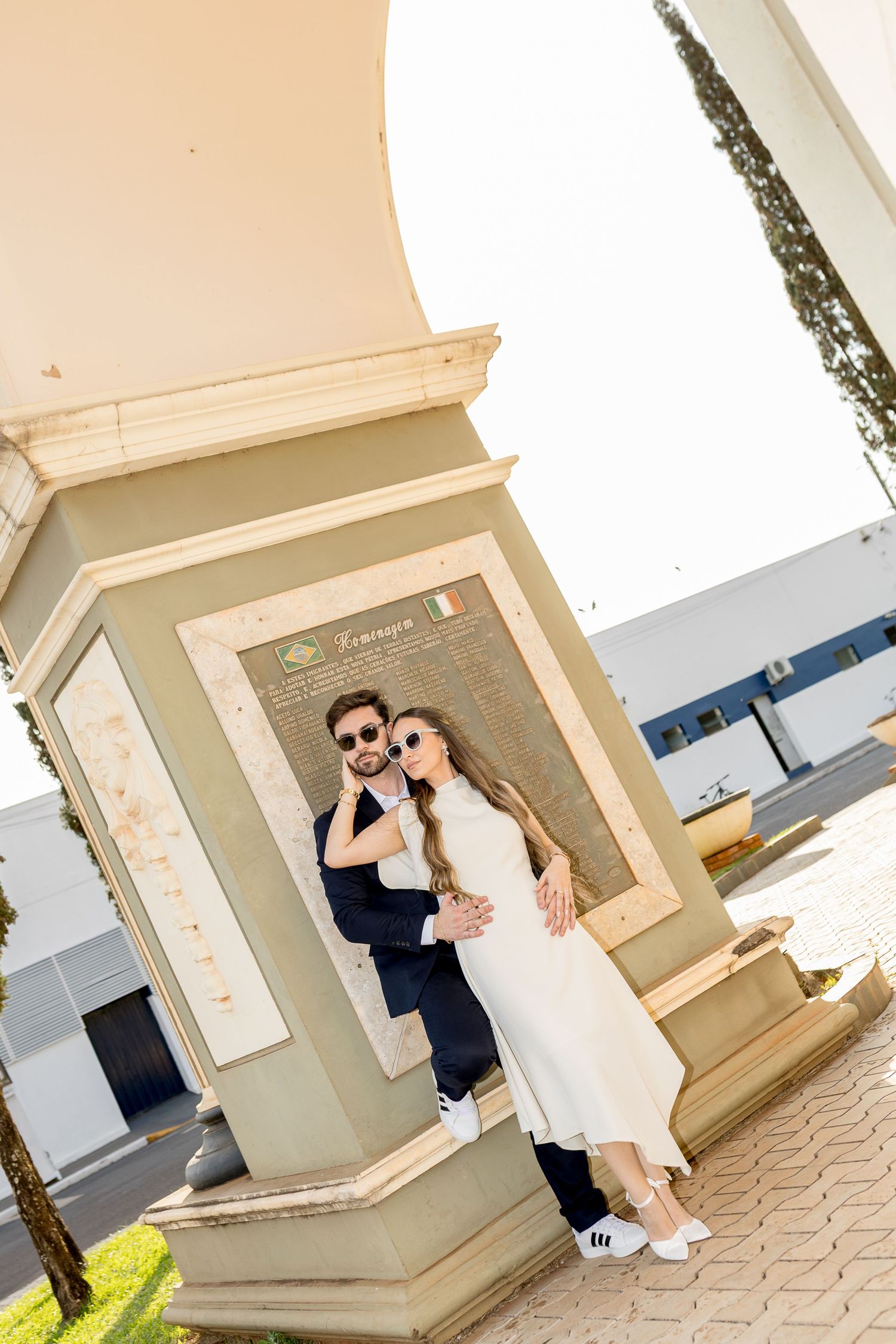 Elegant couple in sunglasses posing together beside an Italian monument plaque in a sunny plaza.