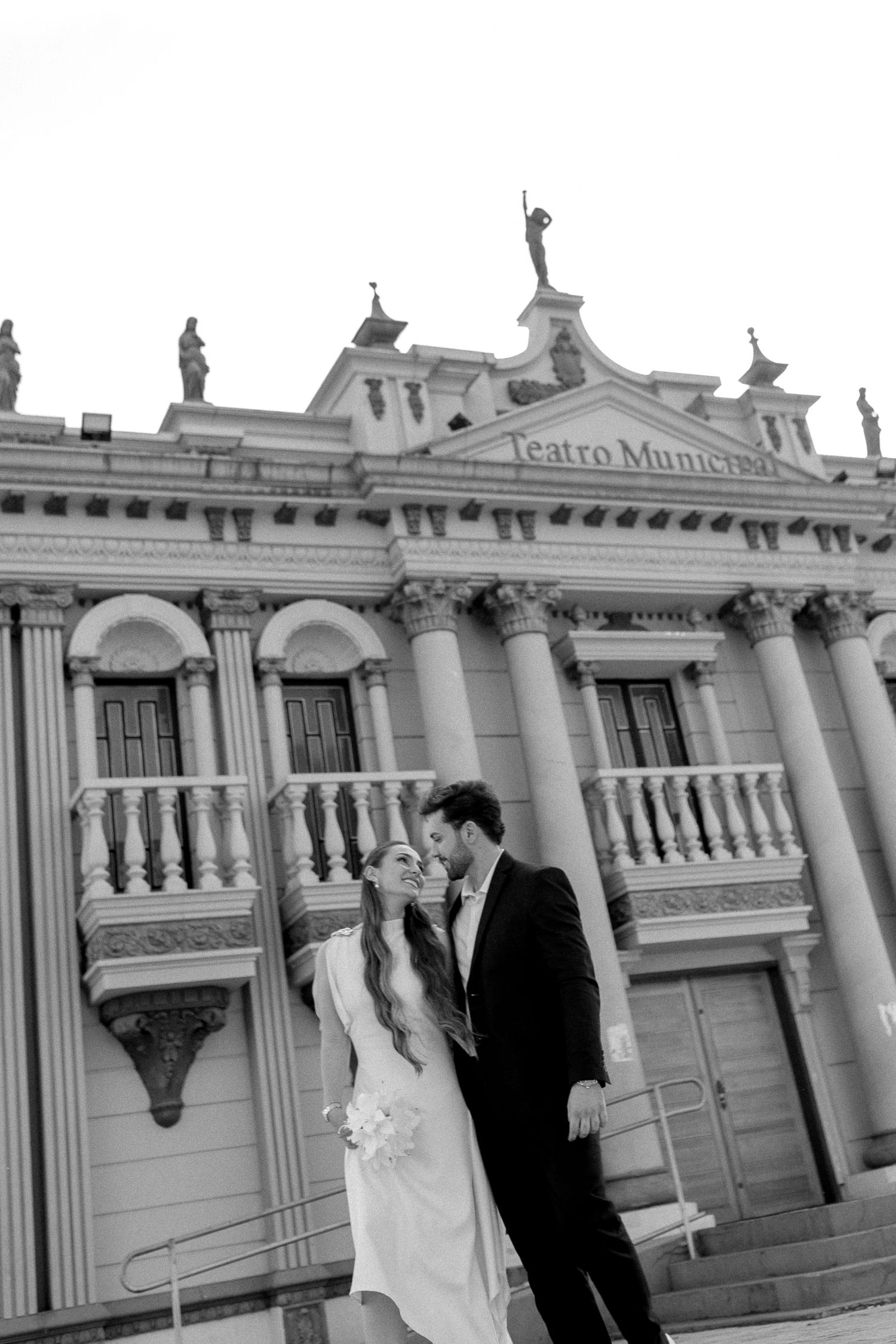 Black and white photo of a wedding couple kissing in front of Teatro Municipal building with classical architecture.