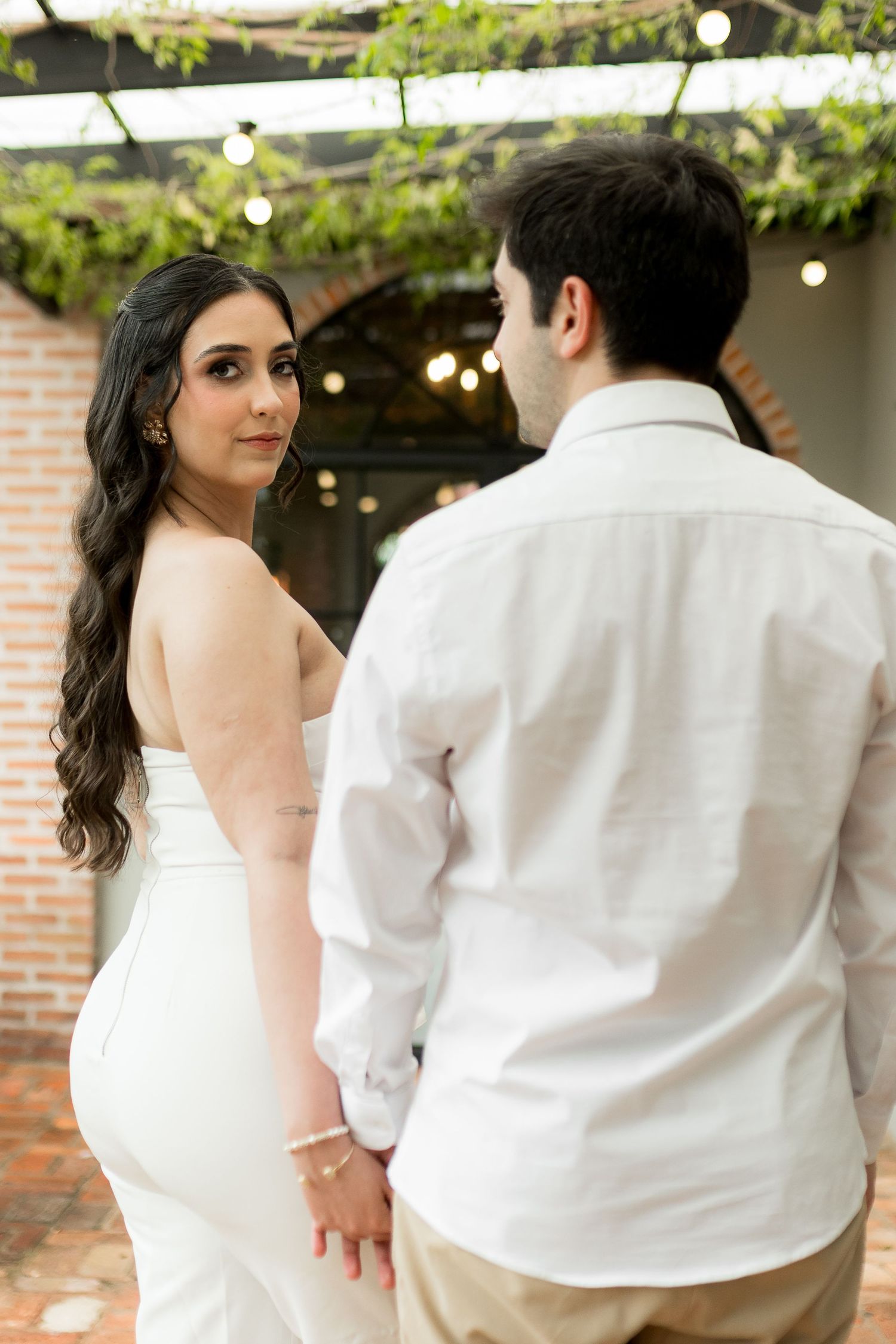 A bride in a white strapless gown looks back over her shoulder while holding hands with her groom outdoors.