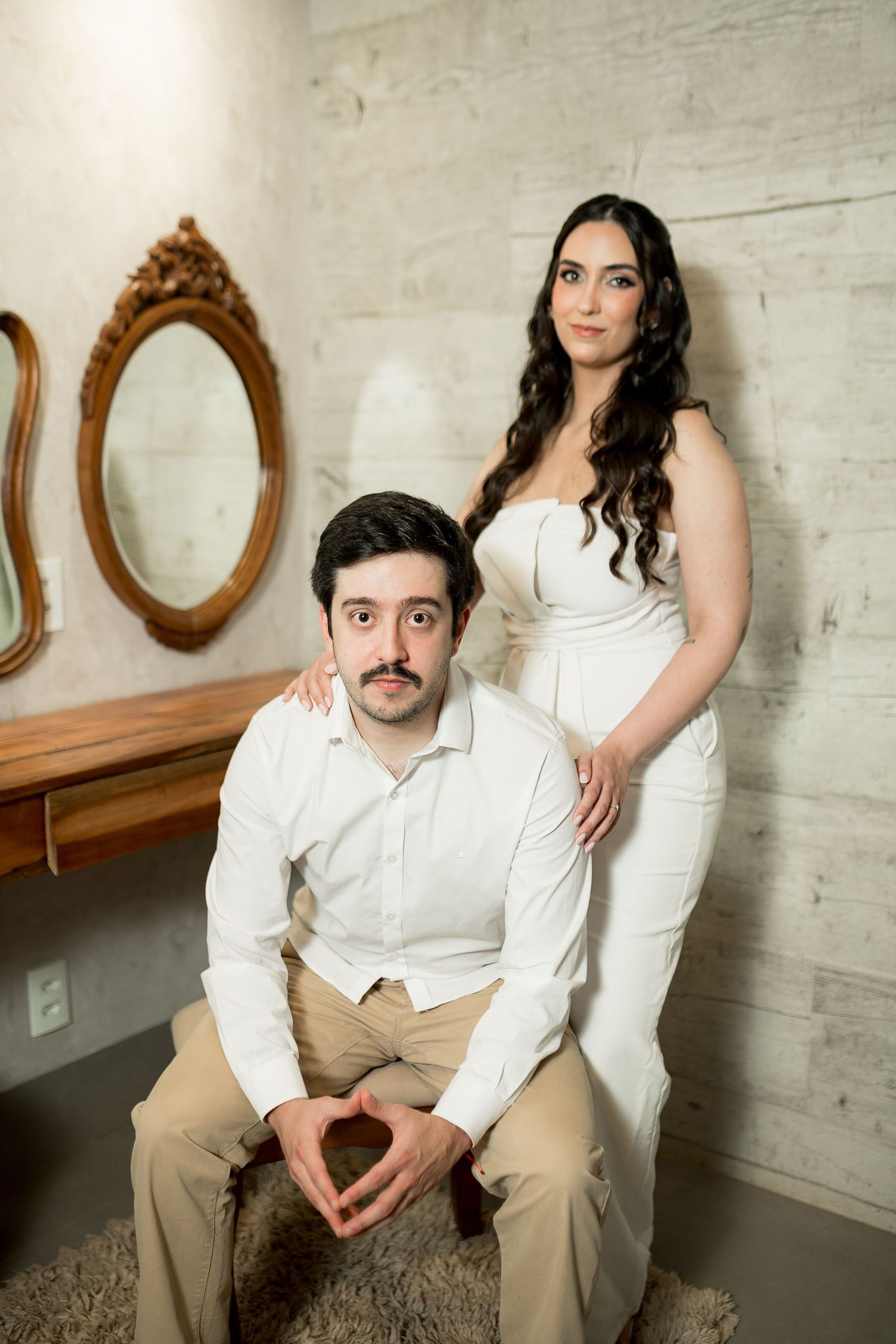 A couple in white outfits poses elegantly in a room with ornate mirrors, the woman standing behind the seated man.