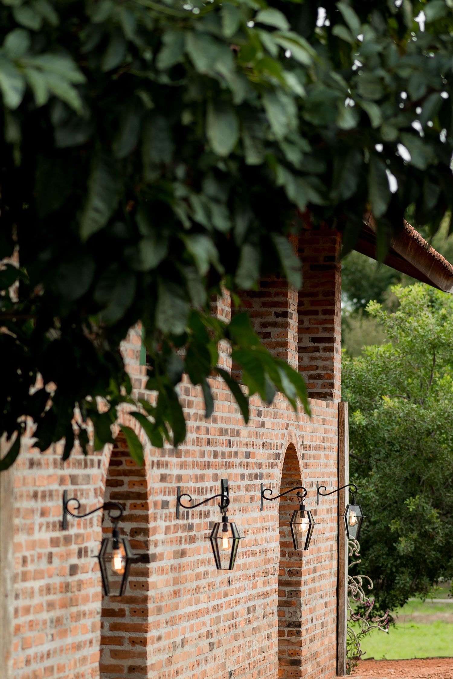 Brick wall with arched niches and decorative iron lanterns, framed by lush green tree branches in an outdoor garden setting.