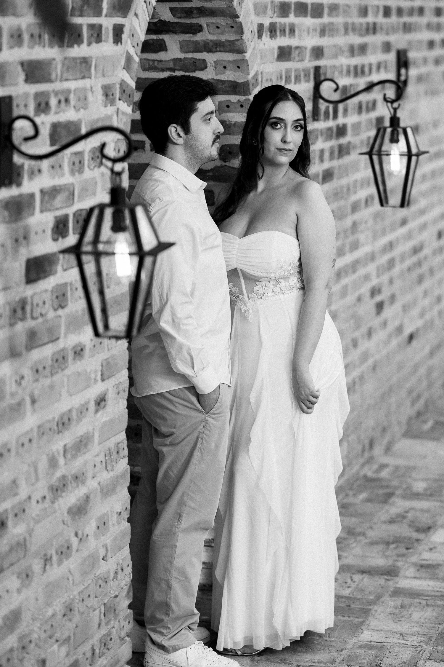 Black and white photo of a couple in formal wear posing against a brick wall with vintage lanterns.