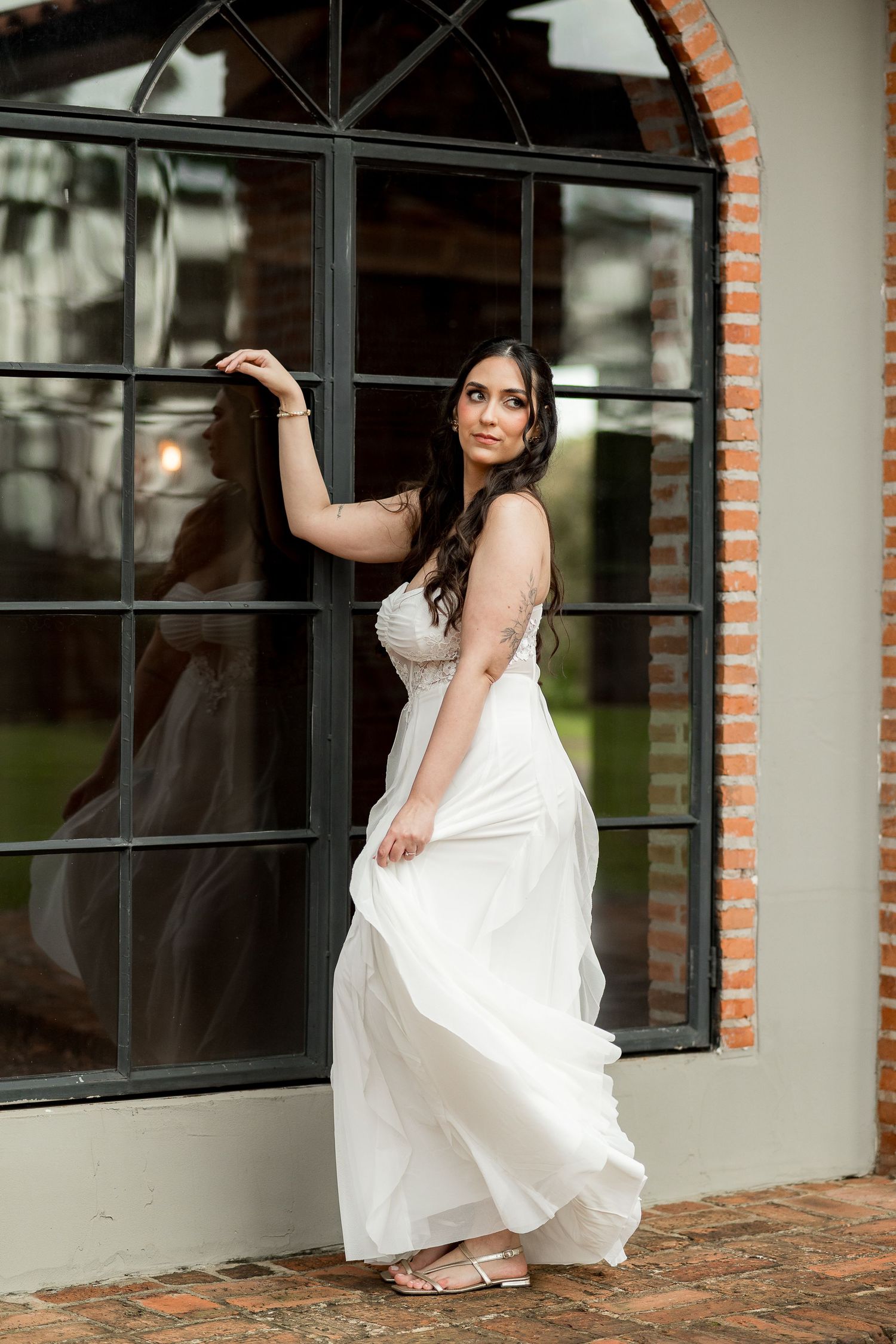 Bride in white lace gown posing by arched window with brick accents at elegant wedding venue.