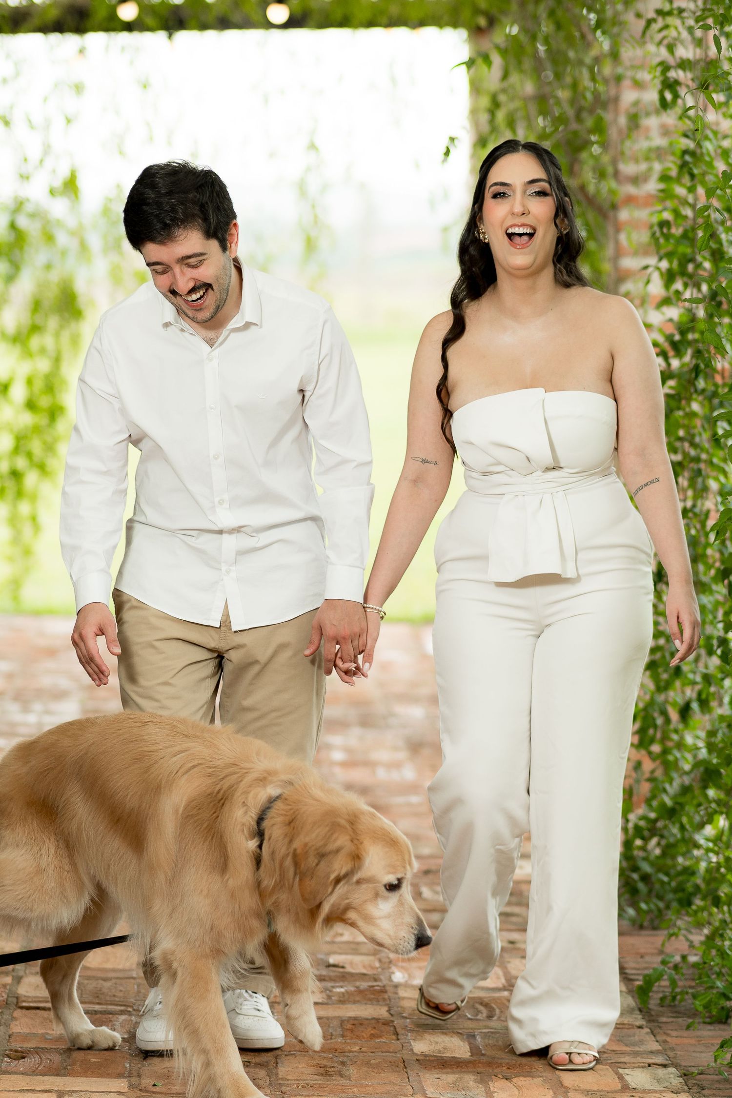 A smiling couple in white outfits holds hands while walking with a golden retriever on a brick path outdoors.