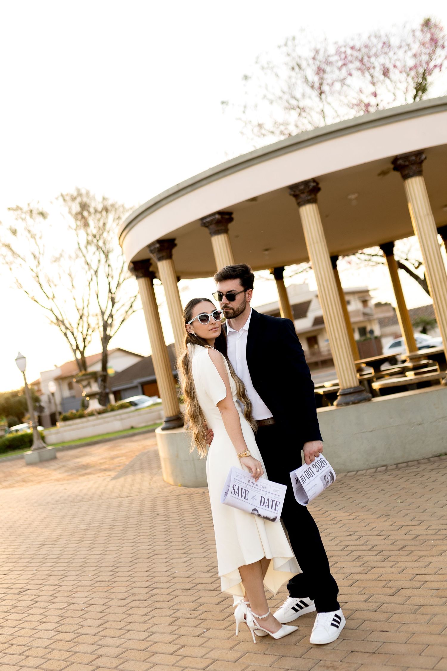Stylish couple in sunglasses holding save the date cards near a columned rotunda at golden hour.