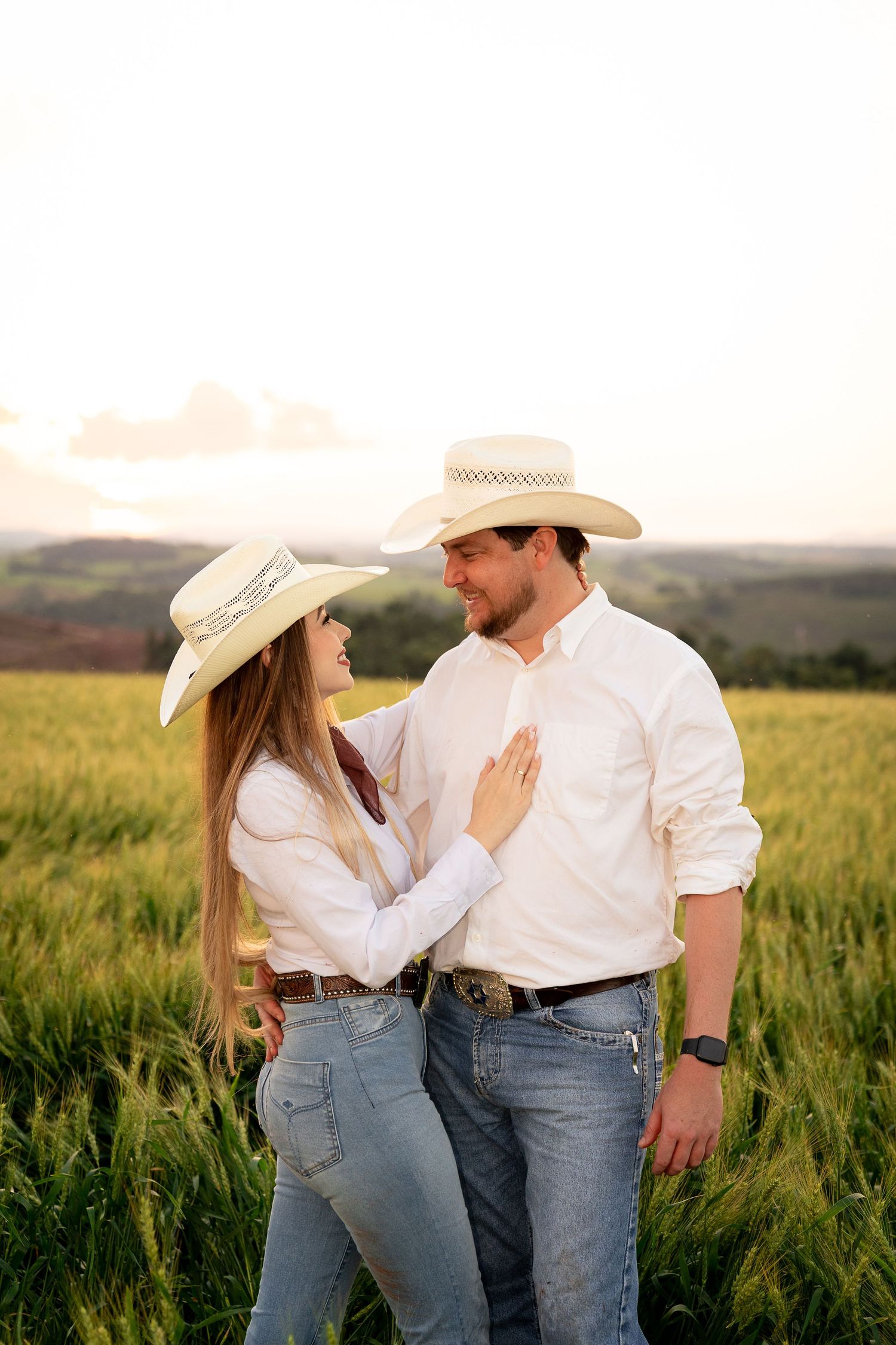 Couple in white shirts and cowboy hats sharing a romantic moment in a lush green field at sunset.