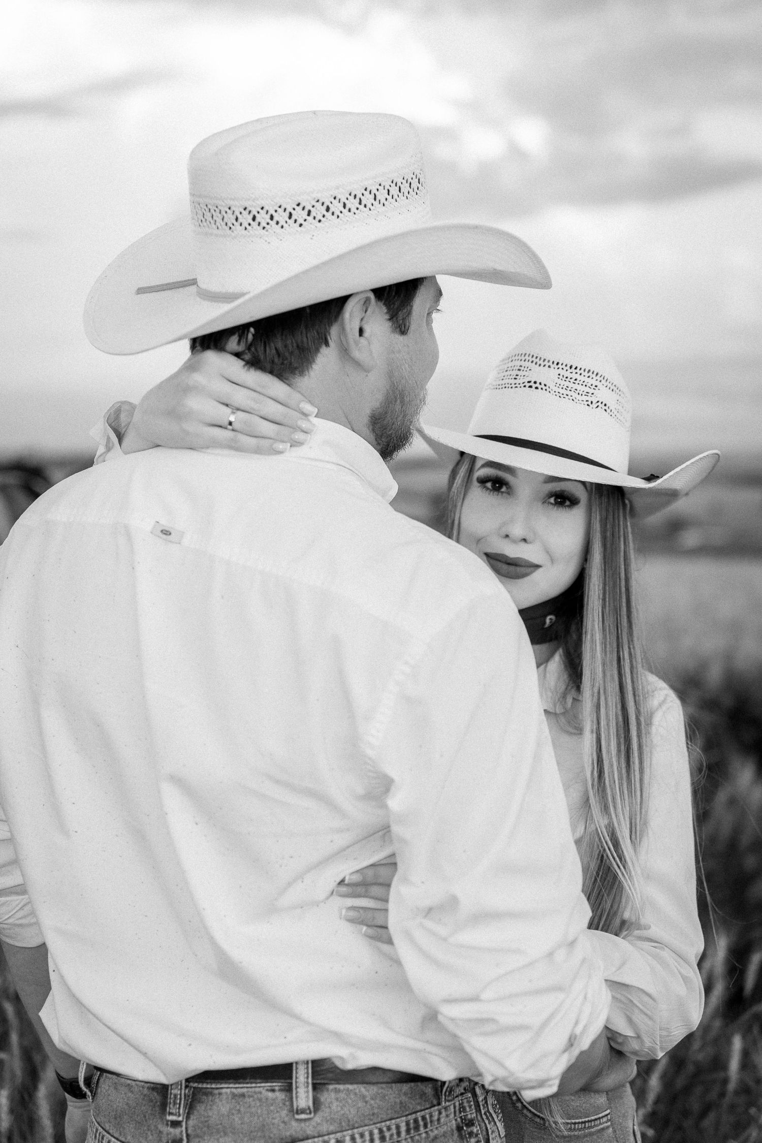 Black and white photo of a couple in cowboy hats embracing in a field, woman smiling at camera.