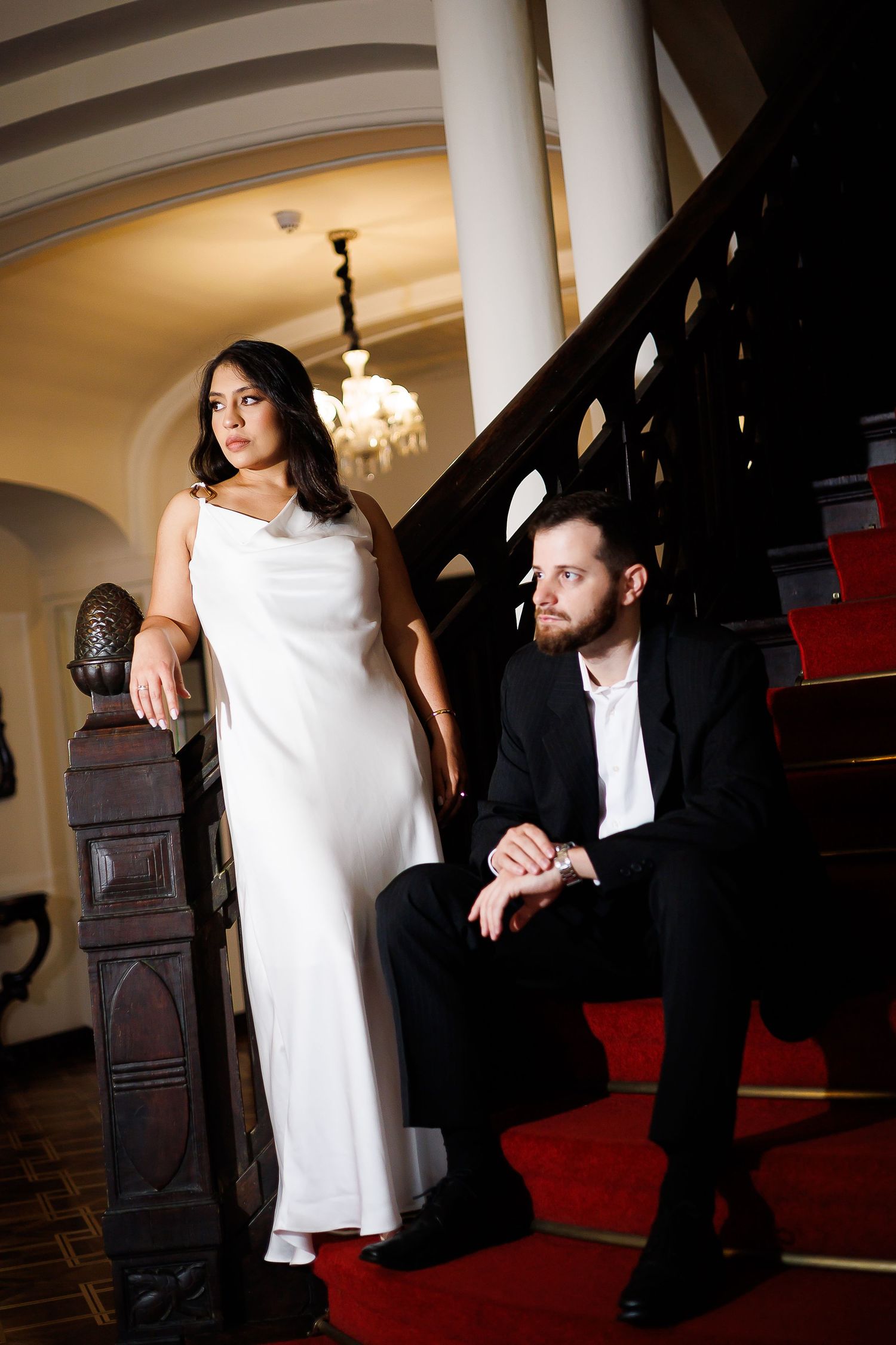 Elegant couple in formal attire posing on red-carpeted stairs in a grand interior with chandelier.