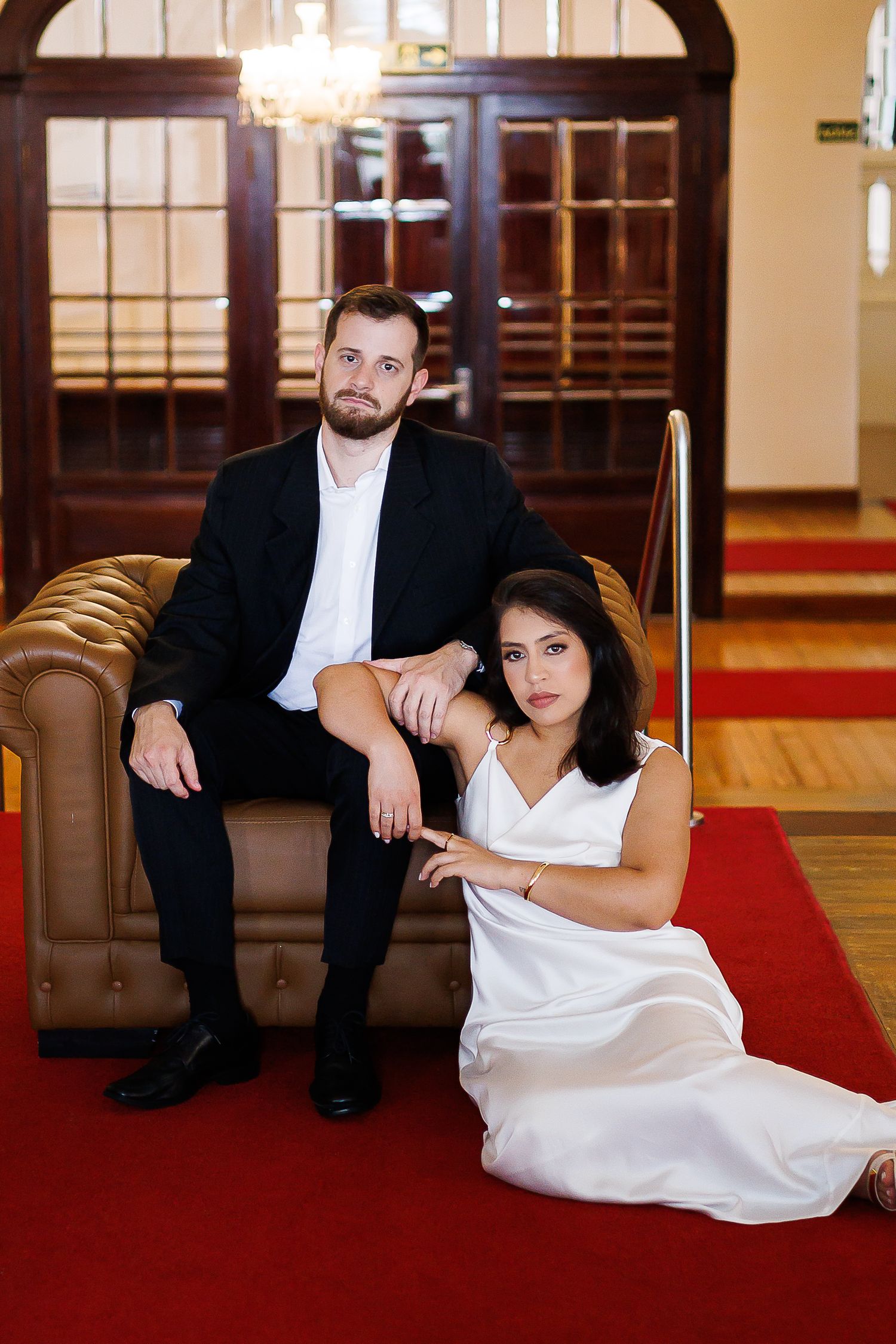 Elegant couple posing in a grand interior, man in black suit on leather chair, woman in white dress on red carpet.