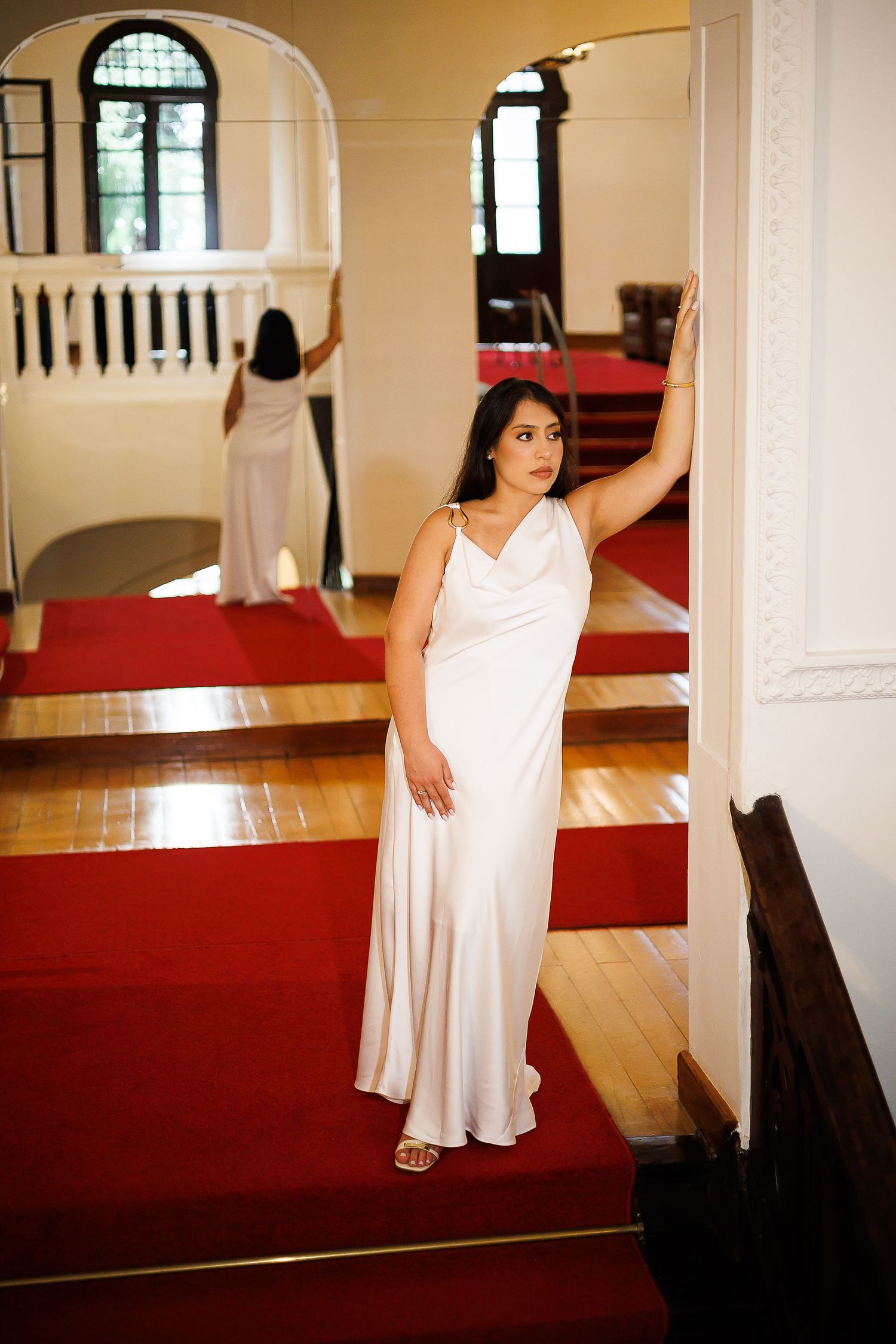 Woman in elegant white satin gown posing in grand hall with red carpet staircase.