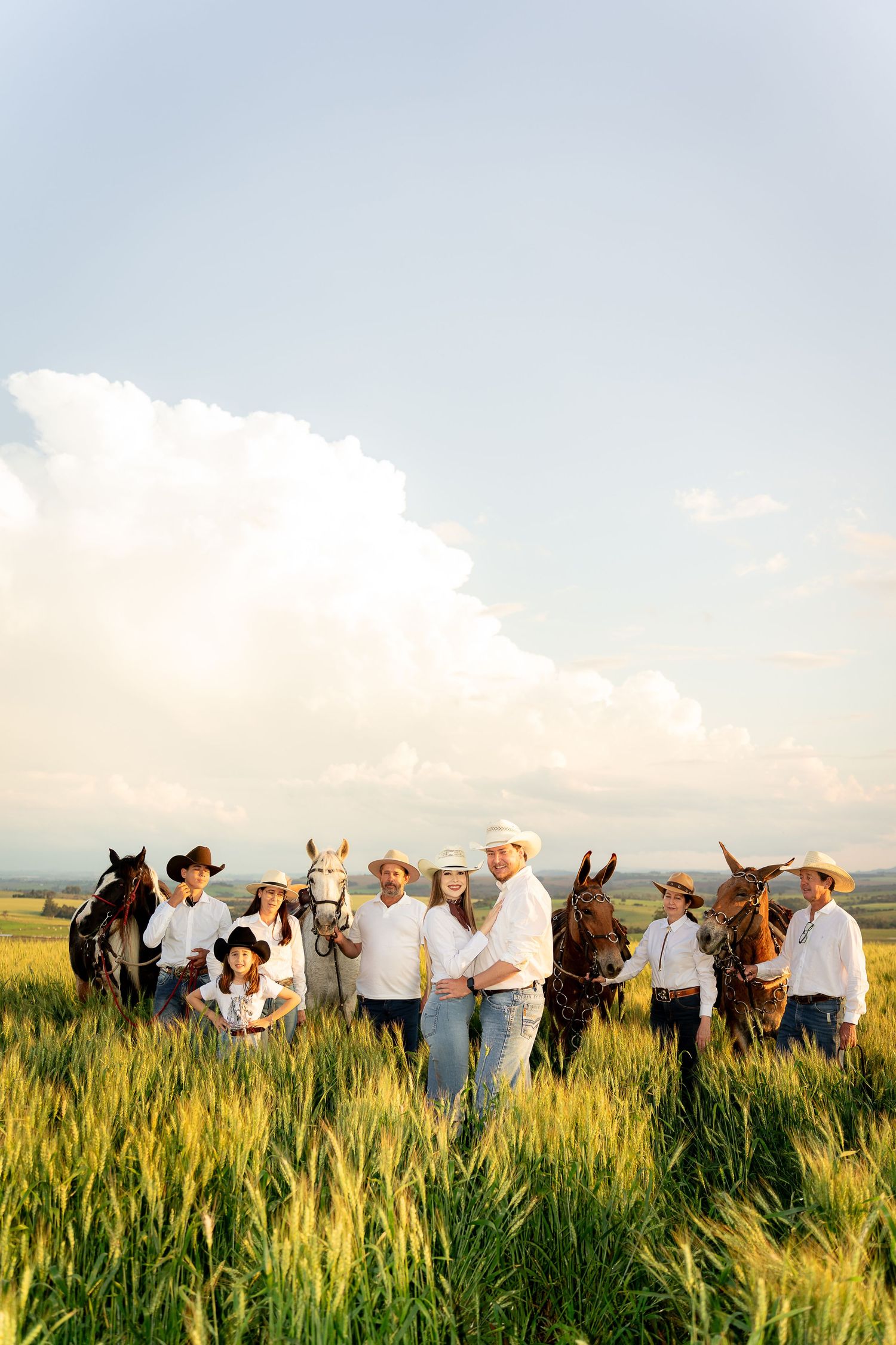A family in white shirts and cowboy hats poses with horses in a green field under a cloudy sky.