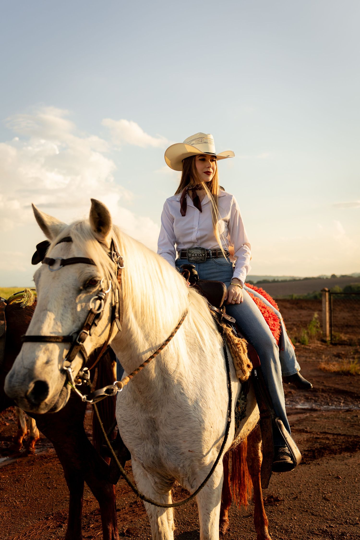 Young woman in white cowboy hat and jeans riding a white horse at golden hour on a rural ranch.