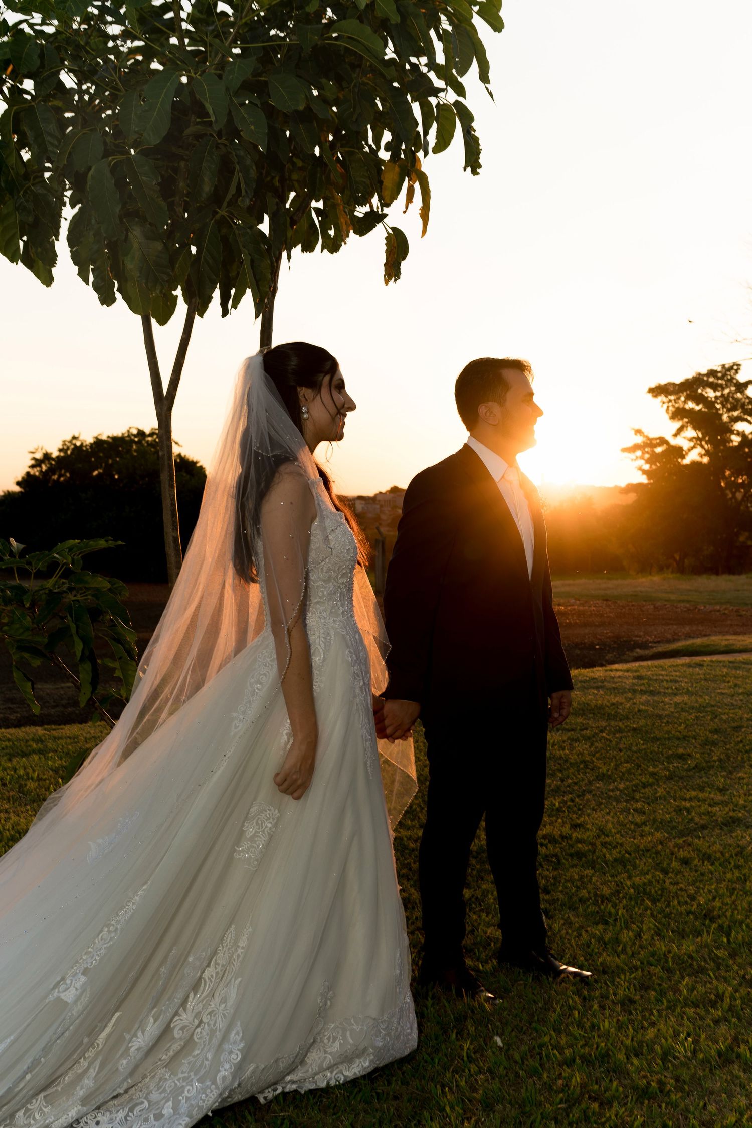 Bride and groom holding hands at golden hour sunset, bride in lace gown with veil, groom in black suit.