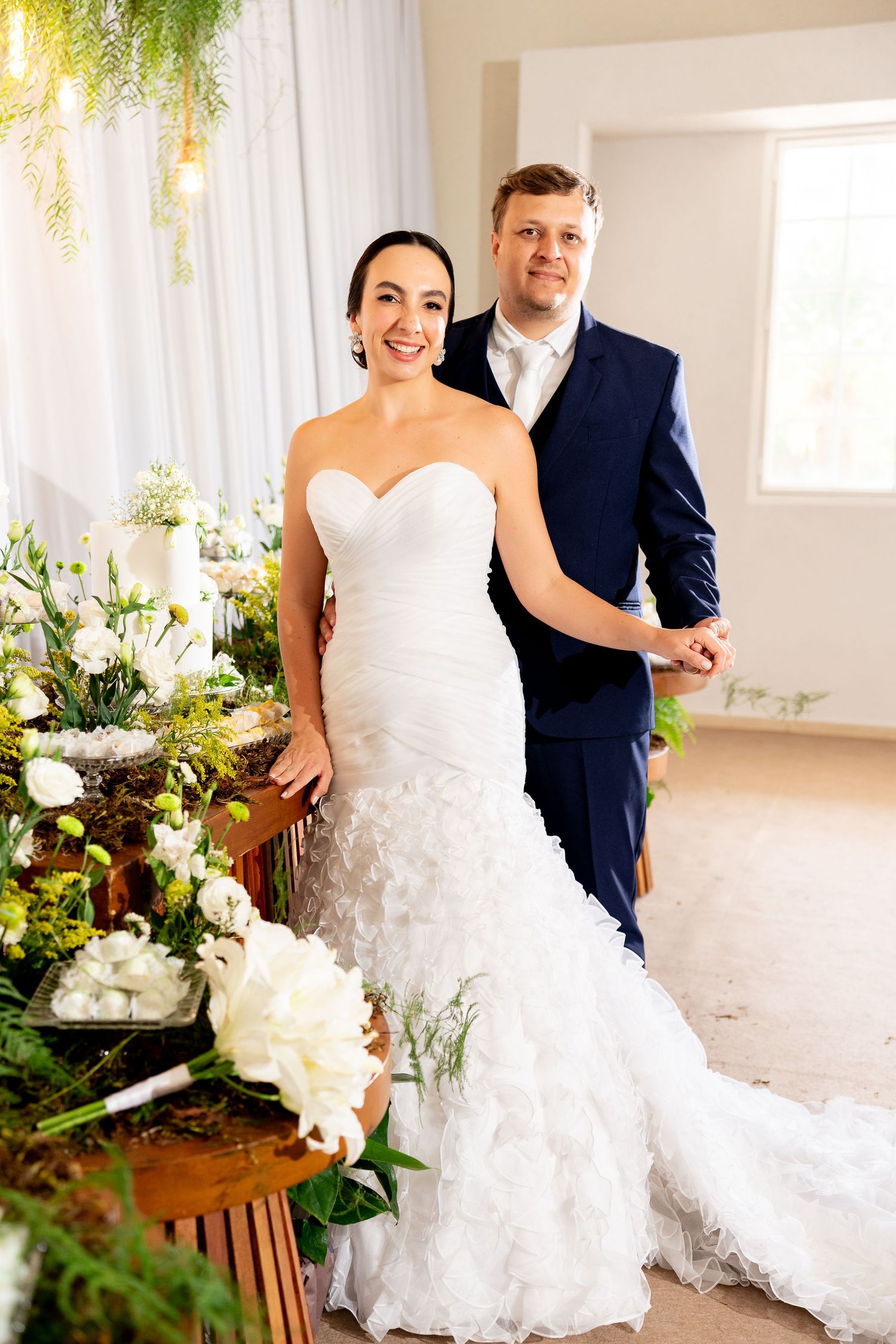 Bride in white mermaid gown and groom in navy suit pose together at elegant floral wedding ceremony decor.