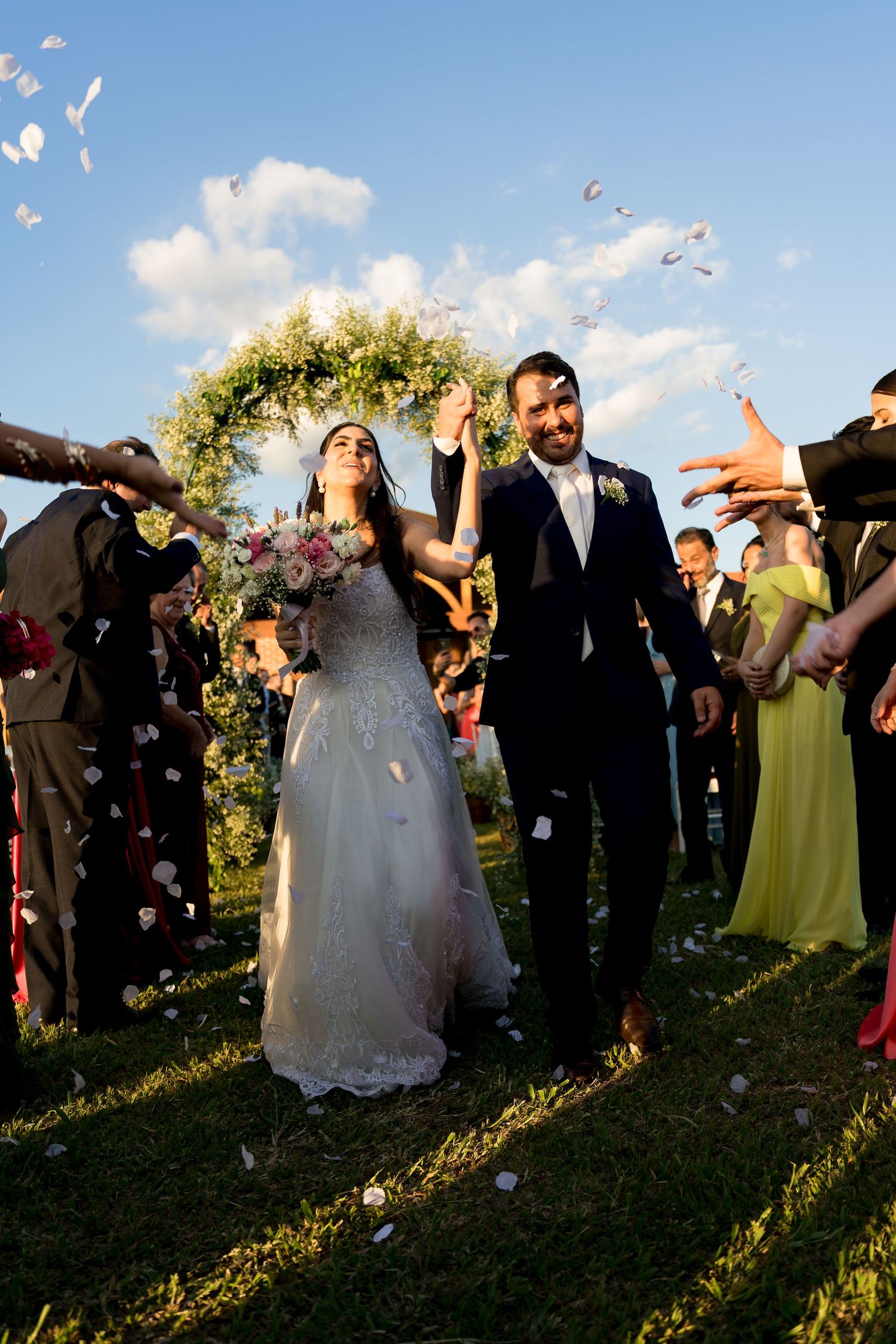 Bride and groom walk joyfully through flower petal confetti shower at outdoor wedding ceremony with floral arch backdrop.