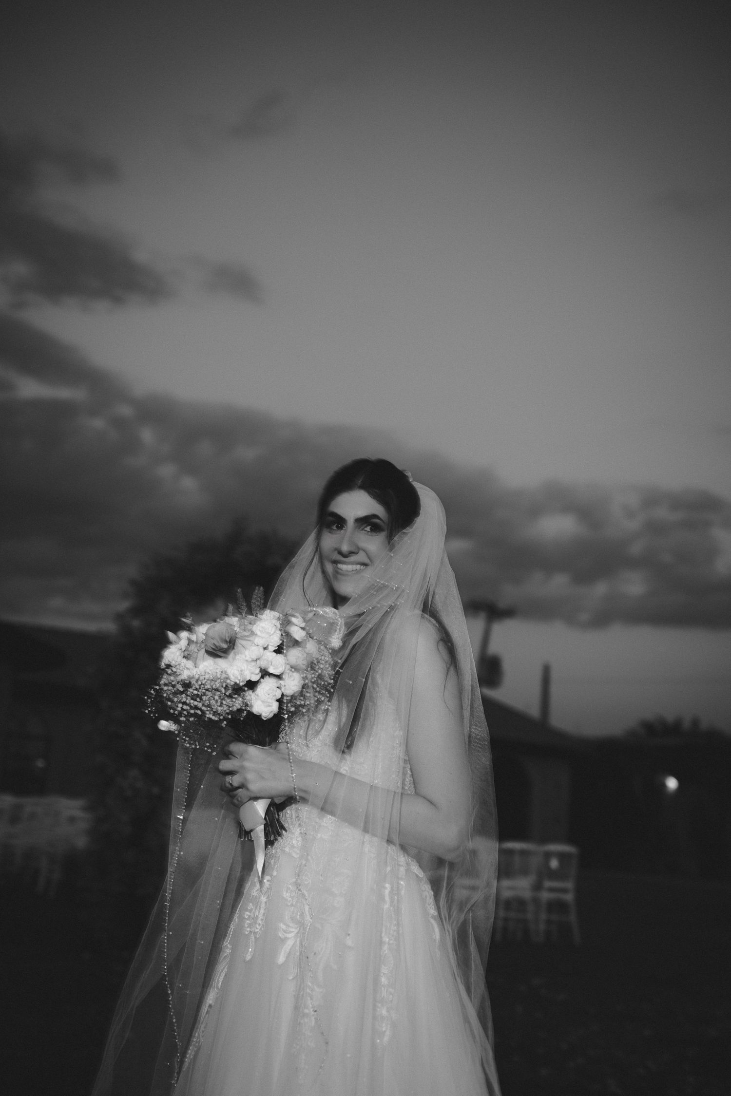 Black and white photo of a smiling bride in a veil holding a bouquet at dusk.