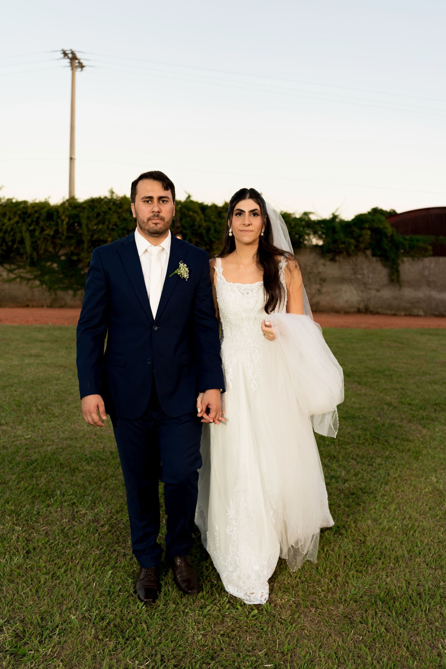 A bride in a white gown and groom in a navy suit hold hands on a green lawn at dusk during their wedding.