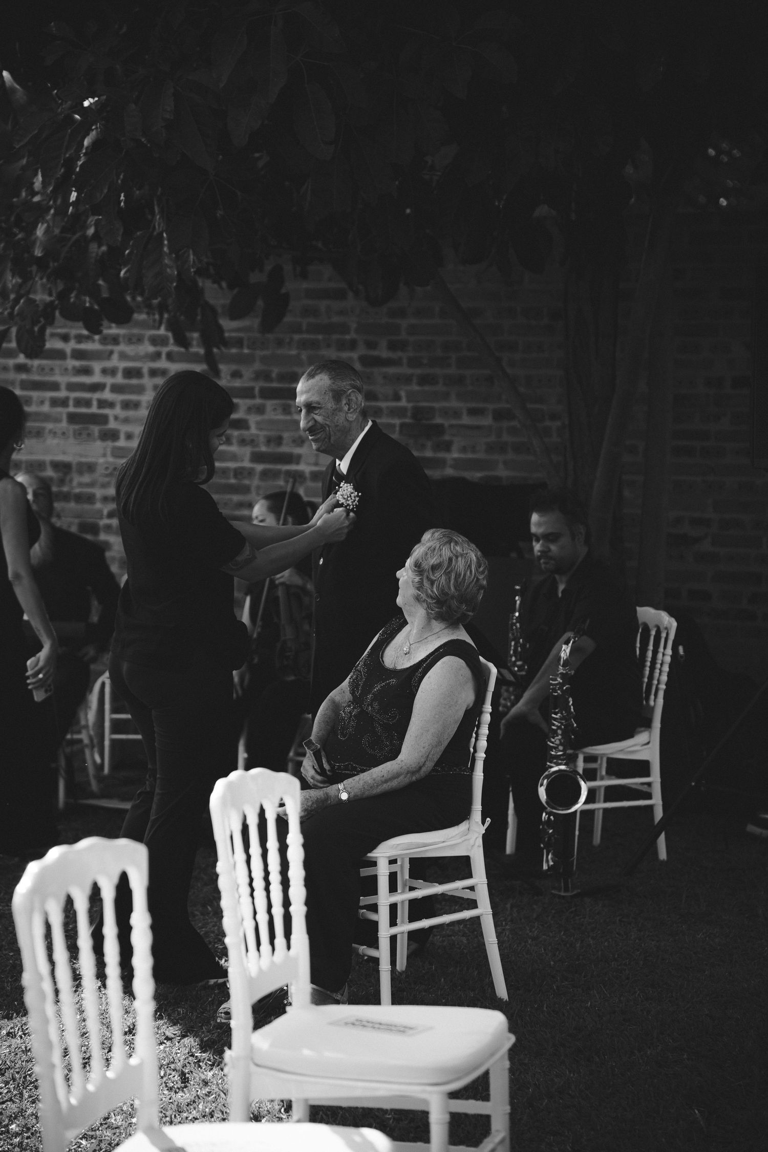 Black and white photo of wedding guests socializing outdoors, with a saxophonist performing near a brick wall.