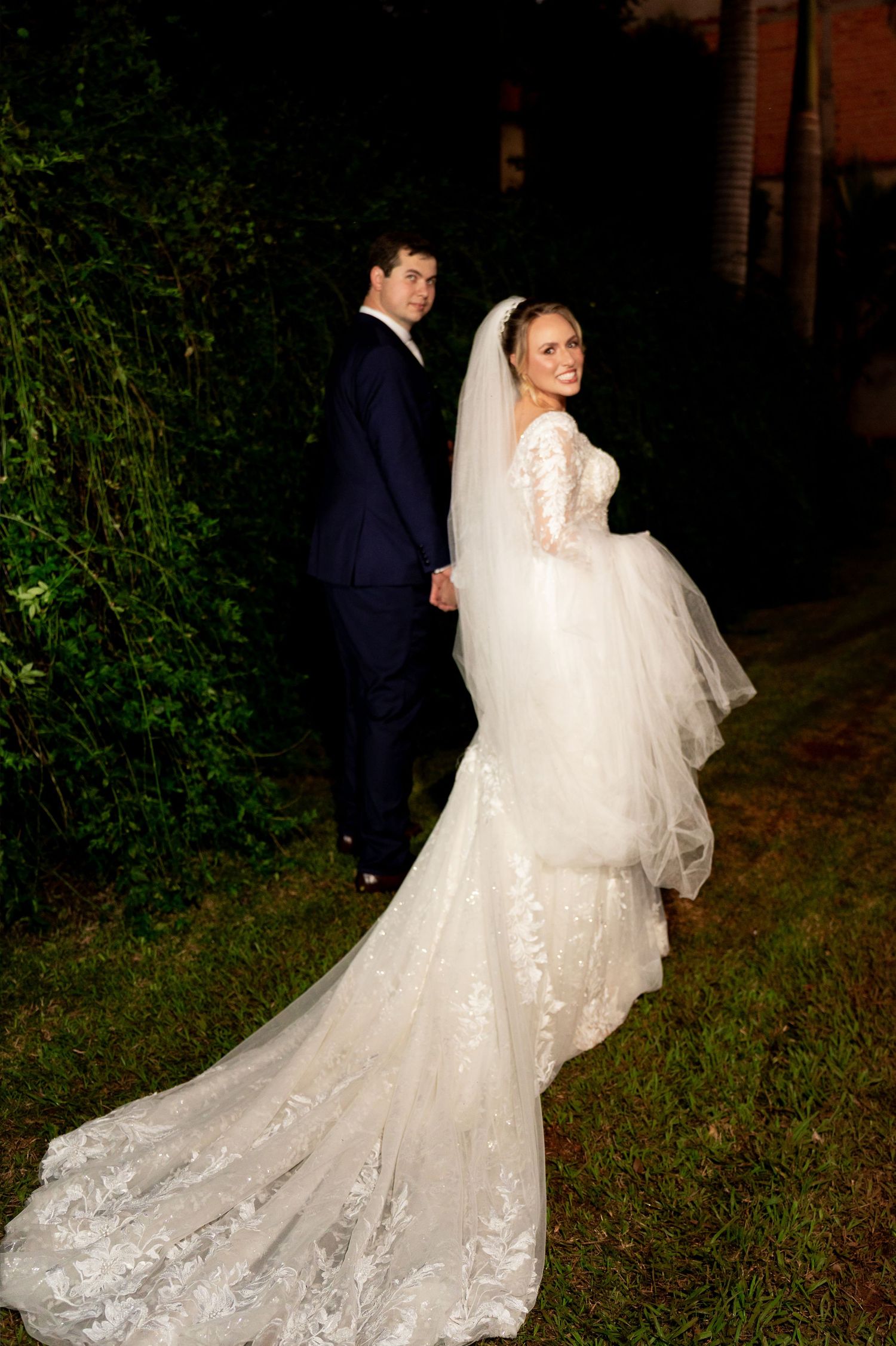 Bride in lace mermaid gown with veil and groom in navy suit pose outdoors at night during wedding portrait.