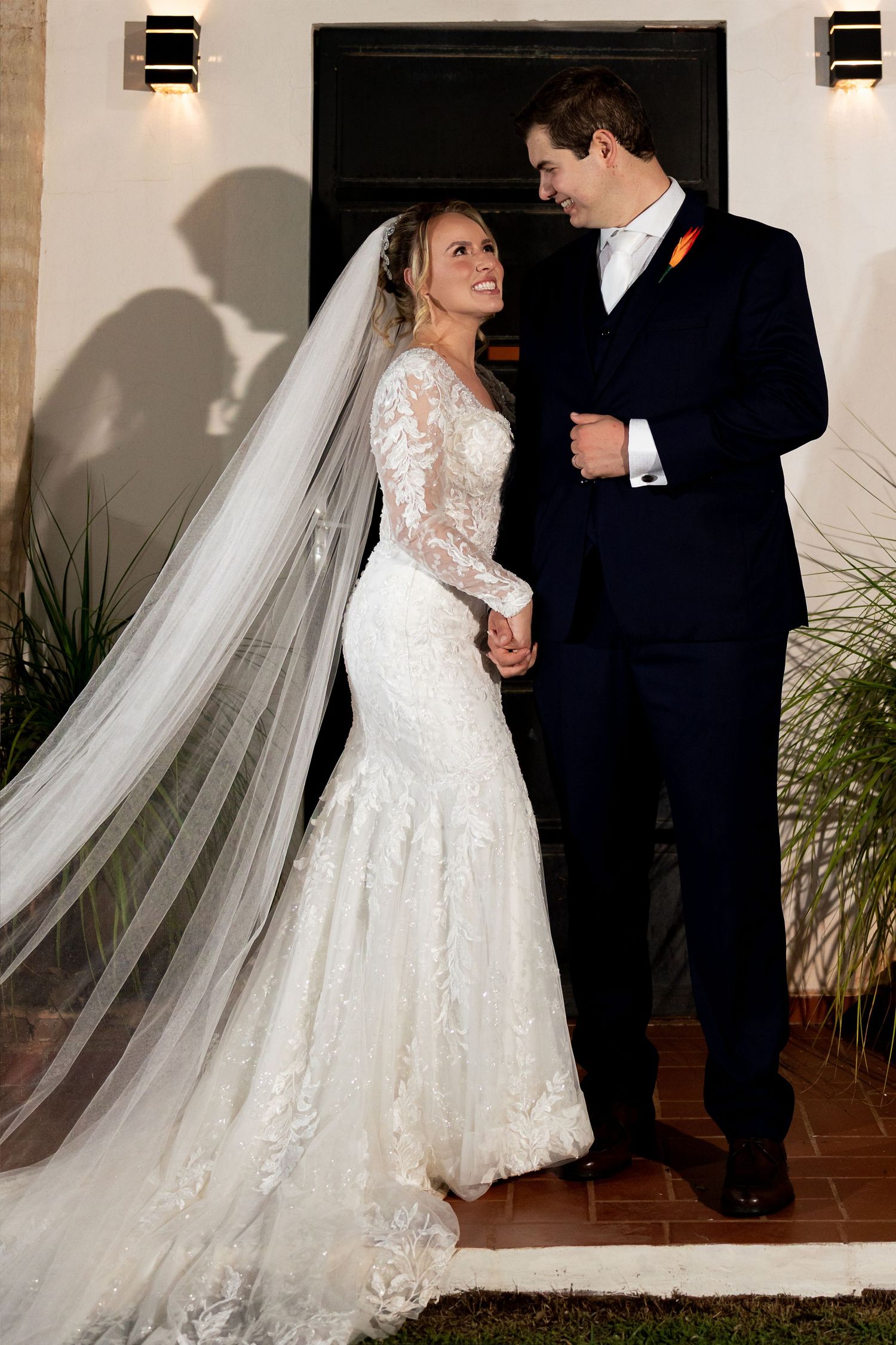 Bride in lace mermaid gown with veil and groom in navy suit share a romantic moment outdoors at night.