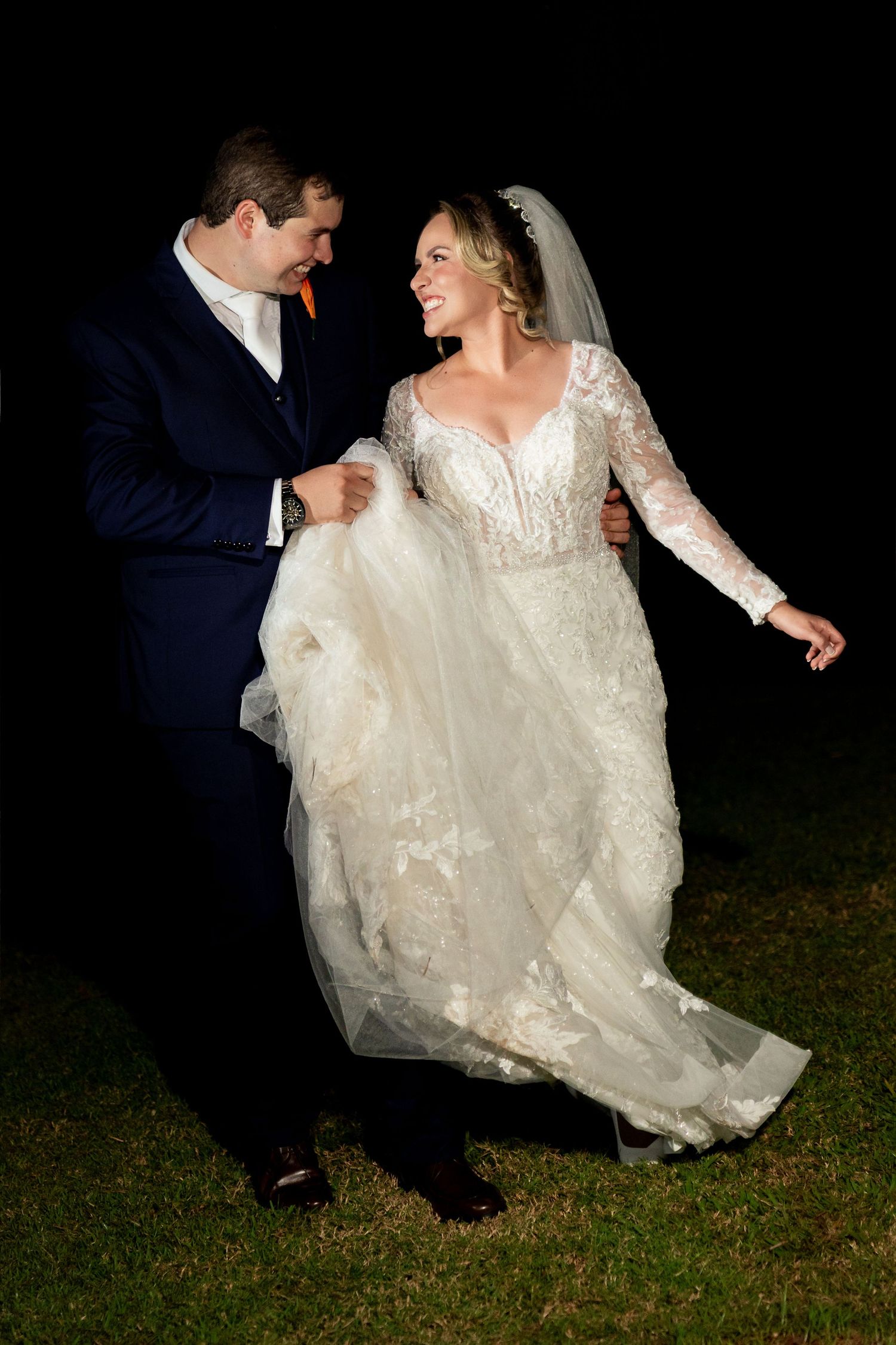 Bride in lace gown and groom in navy suit share a joyful moment outdoors at night during their wedding.