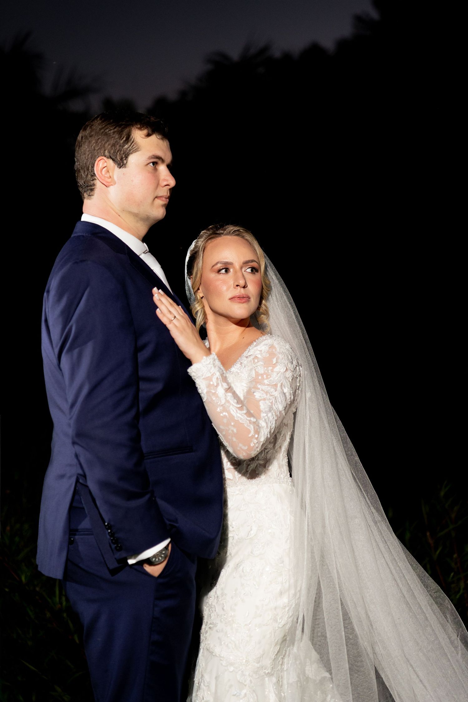 Bride and groom pose elegantly at night, she in lace gown with veil, he in navy suit, against dark backdrop.