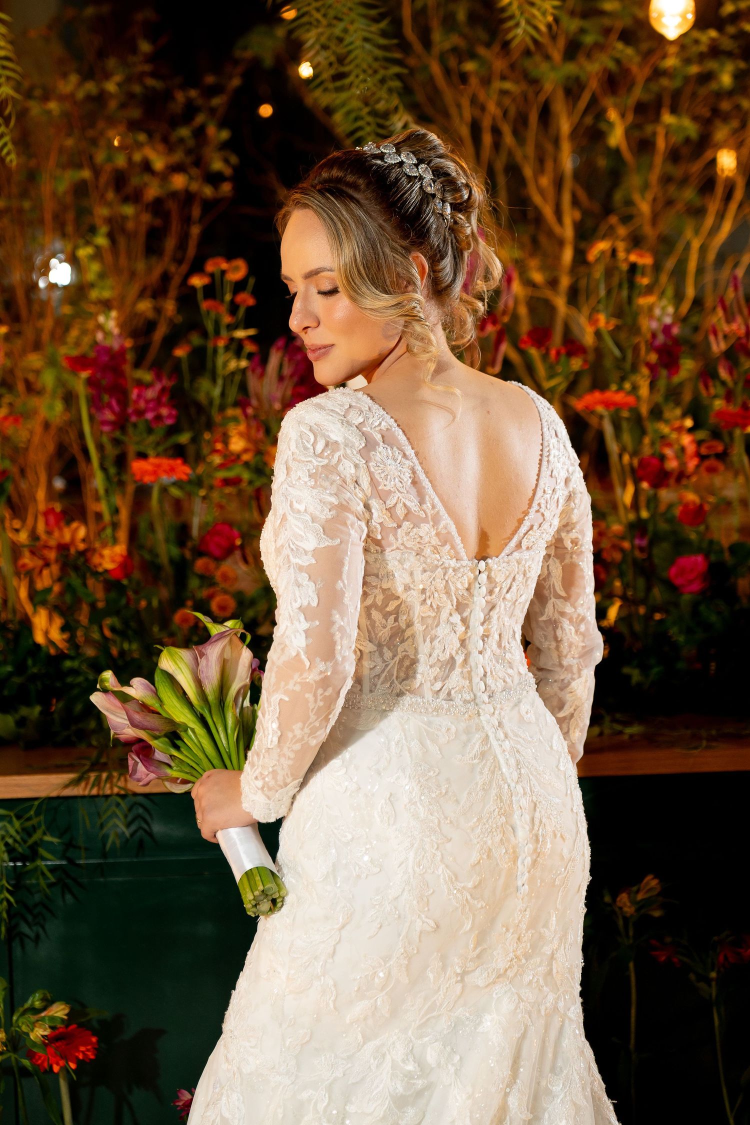 Bride in lace long-sleeve wedding gown with open back, holding bouquet, posed before colorful floral backdrop.